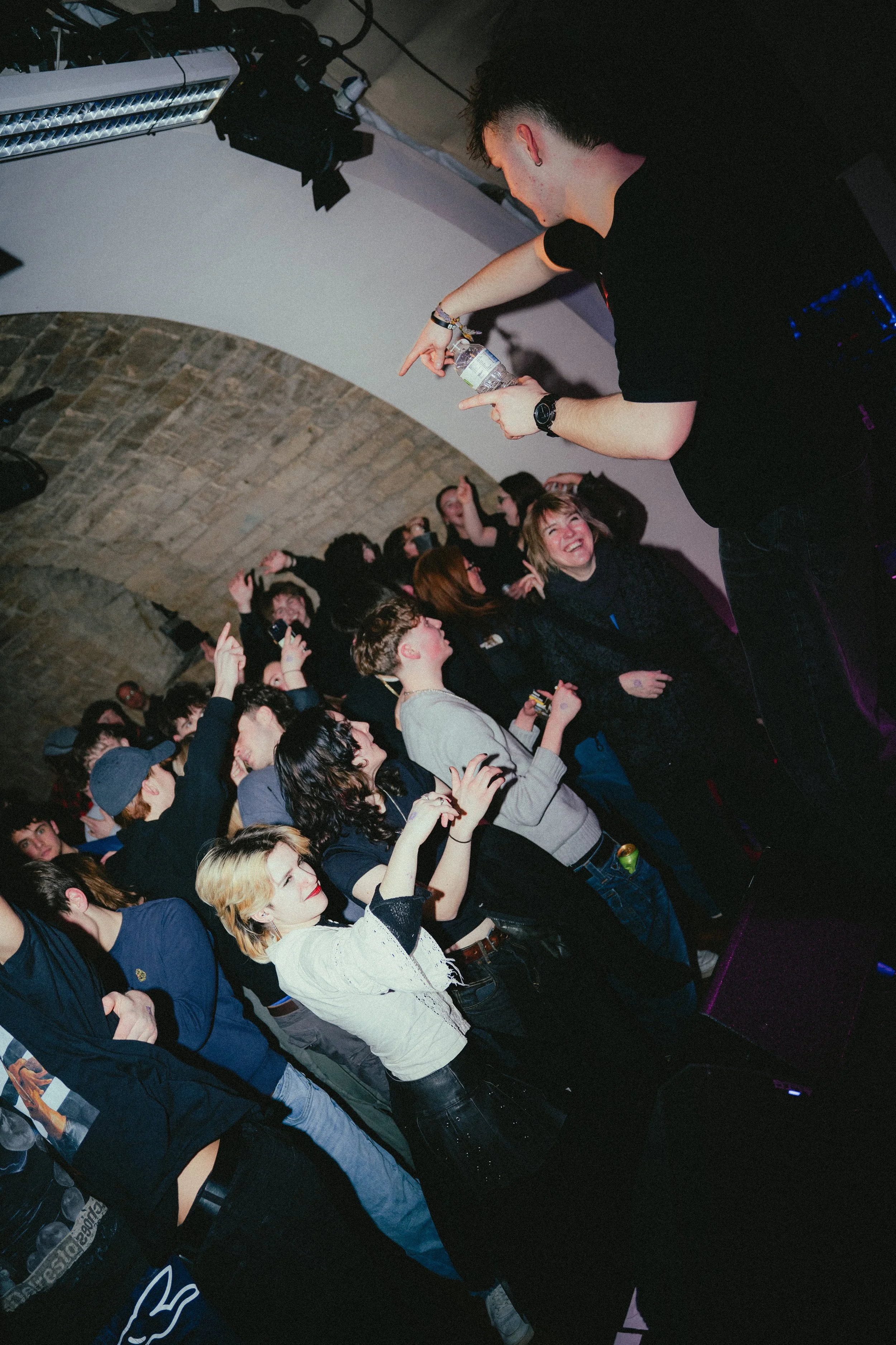 A DJ or performer pointing at a crowd during a live music event in an underground venue, with excited audience members dancing and smiling.