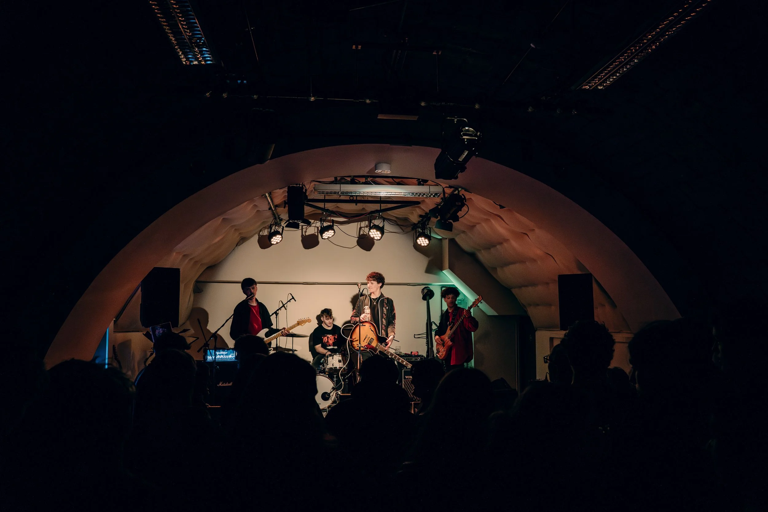 A band performing on stage in a dimly lit venue, with four members playing instruments and one vocalist singing, audience members watching in the foreground.