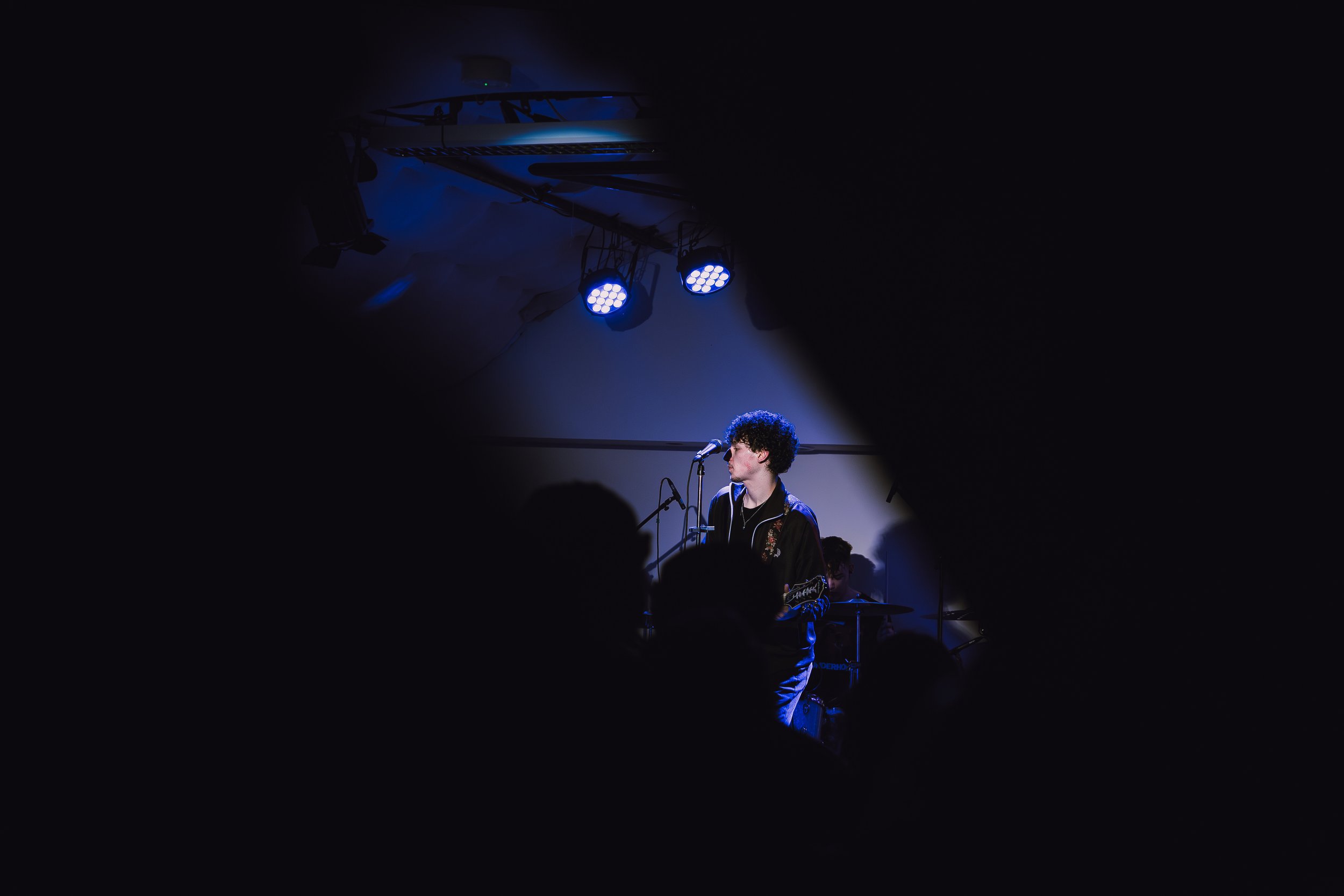 A musician with curly hair playing guitar and singing into a microphone on stage under blue lights, viewed through a gap in the crowd.