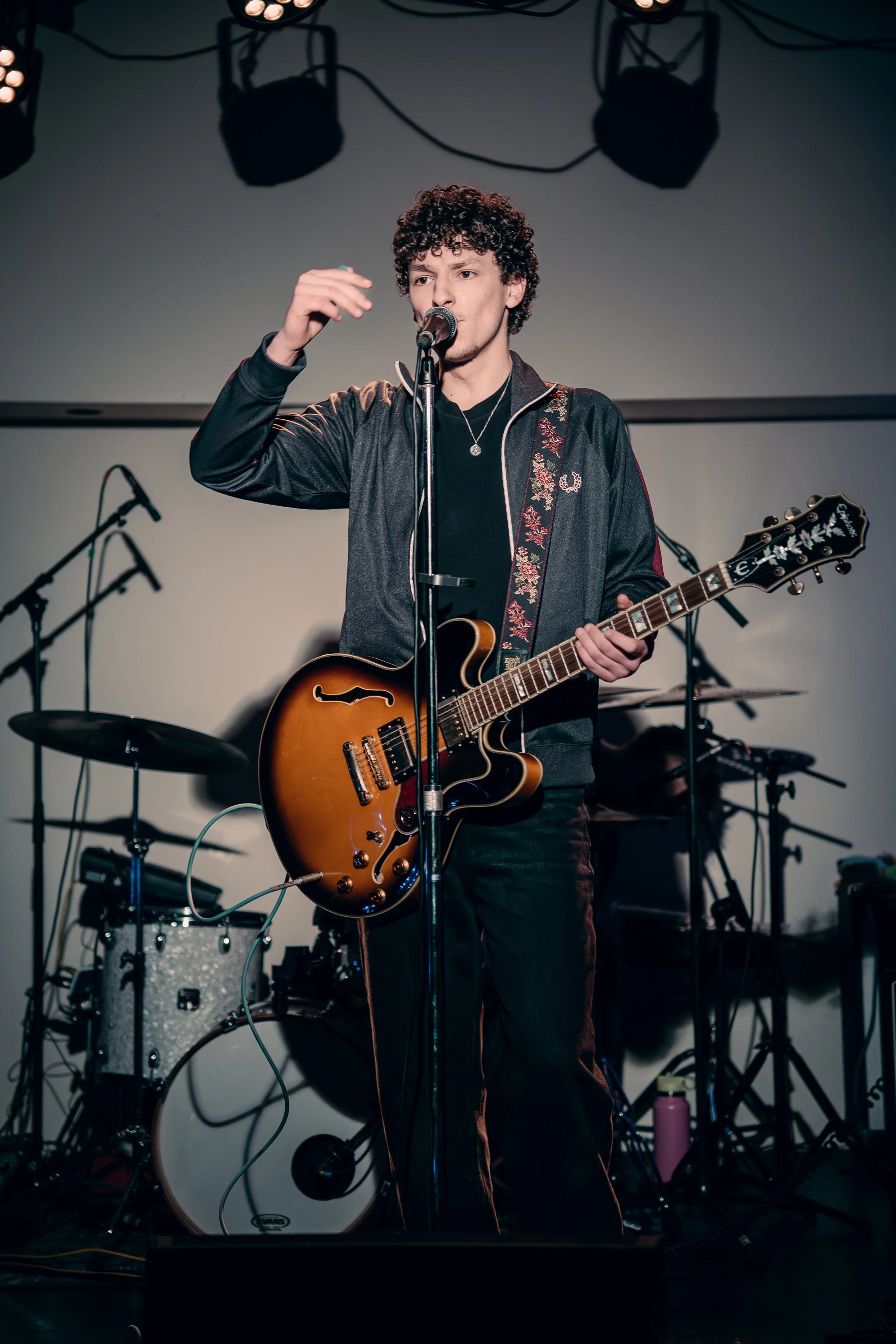 A young man with curly hair performs on stage, singing into a microphone and playing an electric guitar during a live music performance.