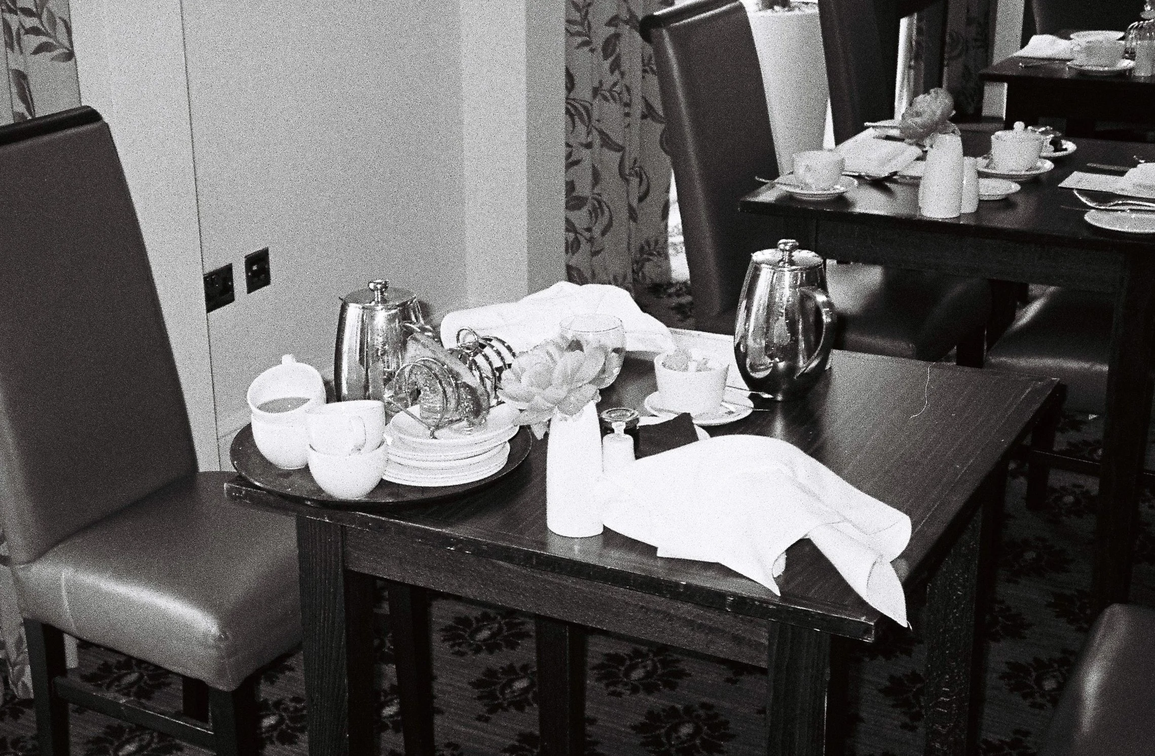 A dining table set for a meal with cups, teapots, plates, and cutlery, with chairs around it, in a room with patterned curtains and a carpet.