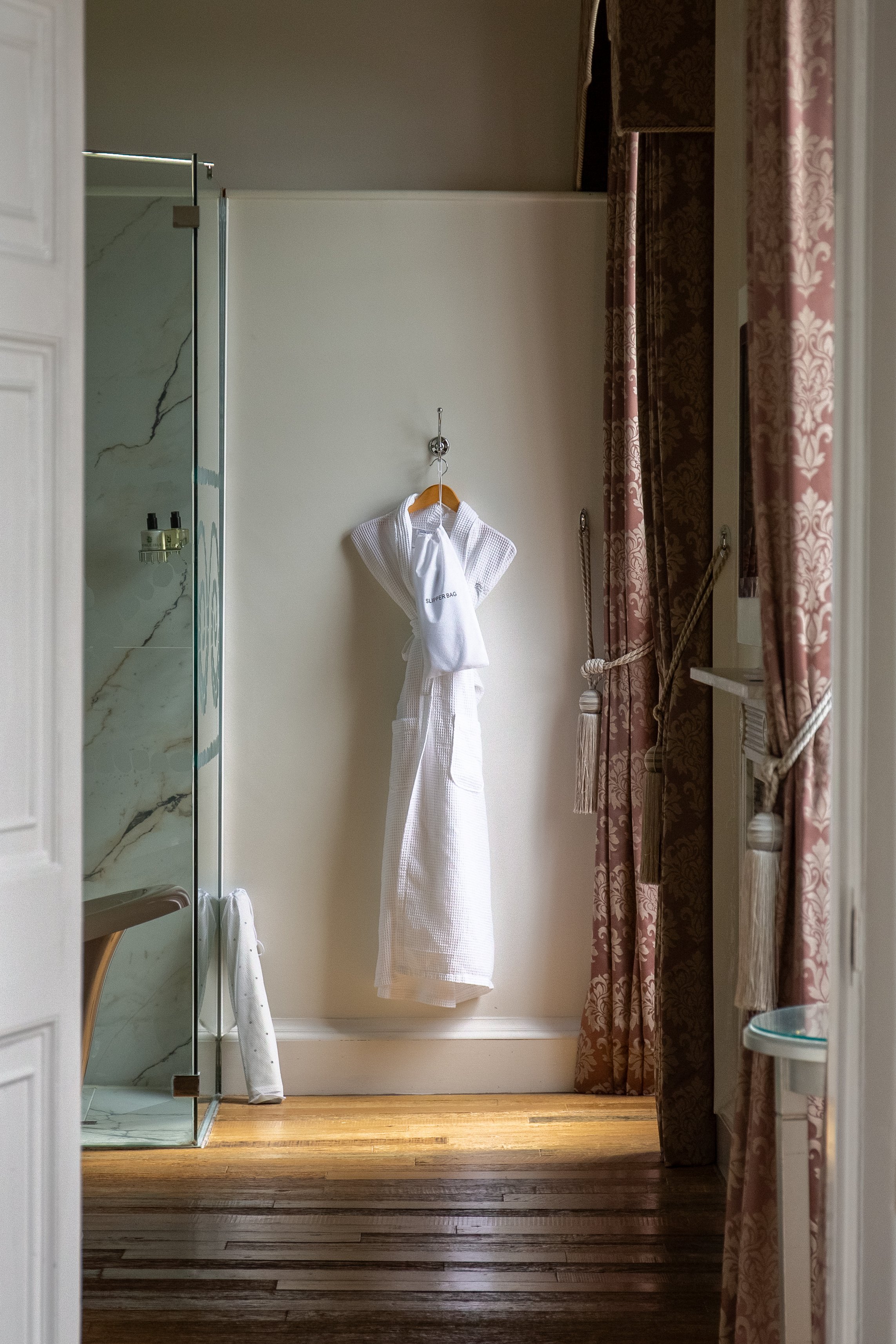 Bathroom with hanging bathrobe, towel, and shower door, with pink curtains and wooden floor.