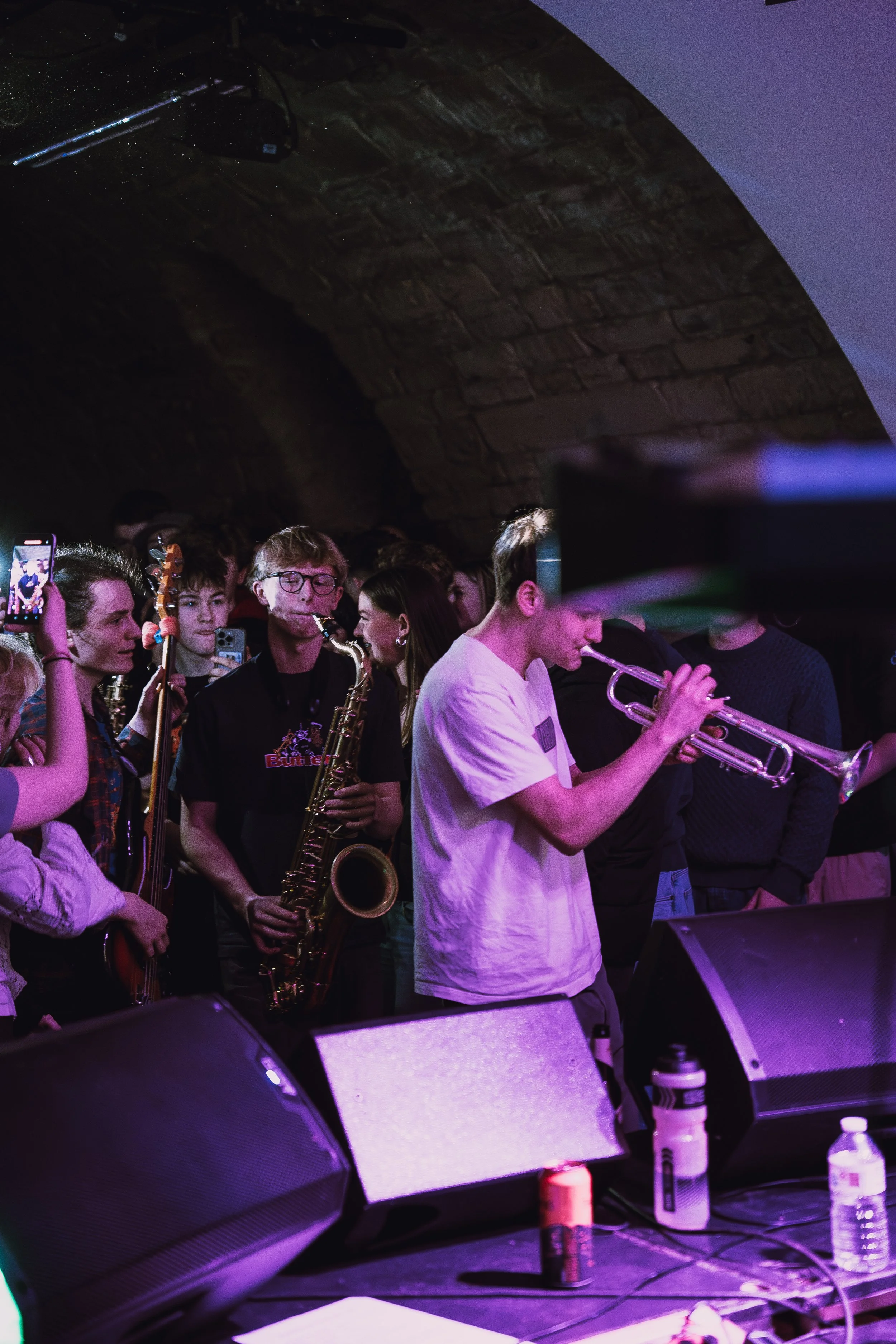 Musicians performing on stage in a venue with a brick arch ceiling, playing saxophone and trumpet, surrounded by audience members.