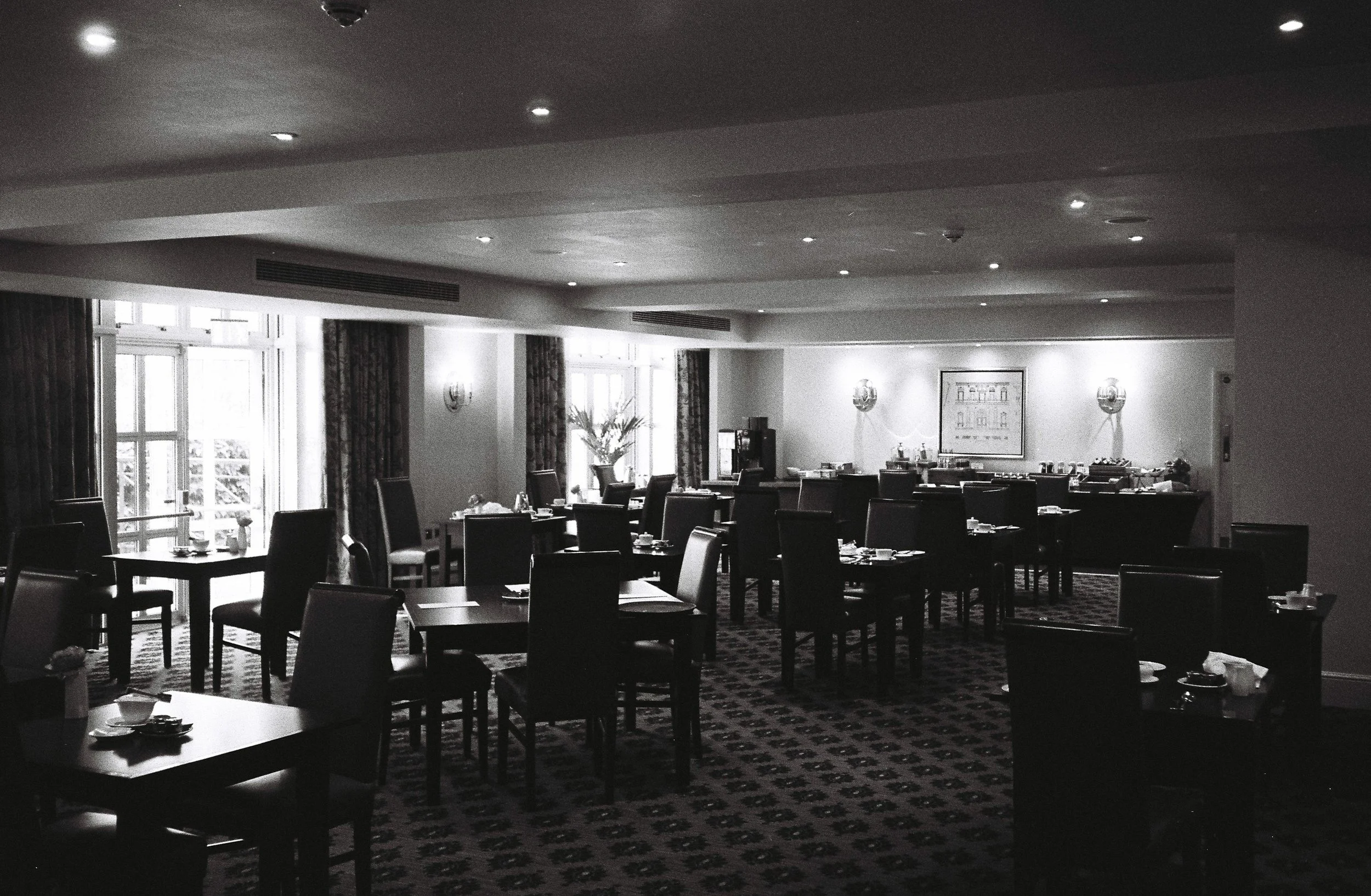 Empty dining room with dark wood tables and chairs, patterned carpet, large windows with curtains, and wall-mounted lights.