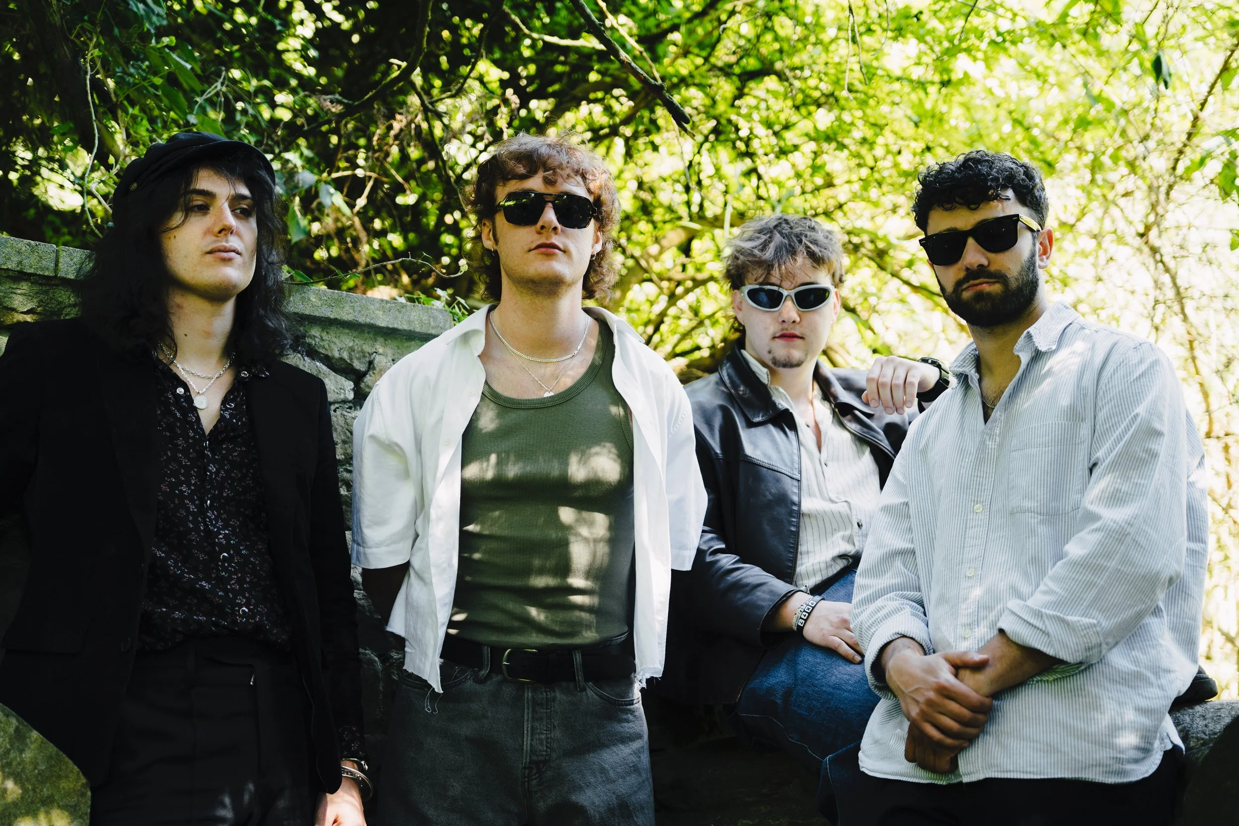 A group of four young men with varying hair styles and sunglasses standing outdoors under a tree with green leaves.