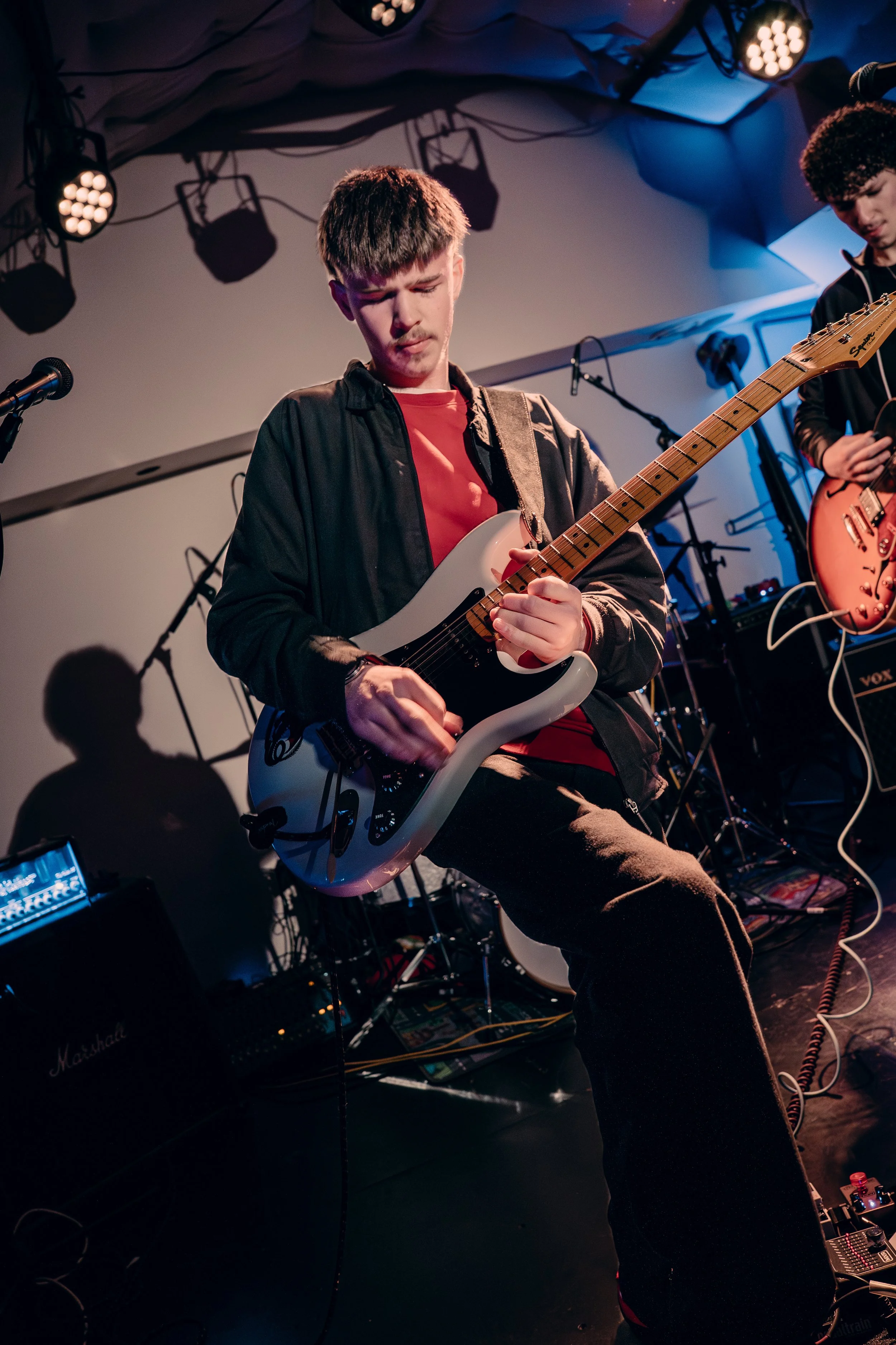 A young man playing an electric guitar on stage during a live music performance, with stage lights overhead and musical equipment in the background.
