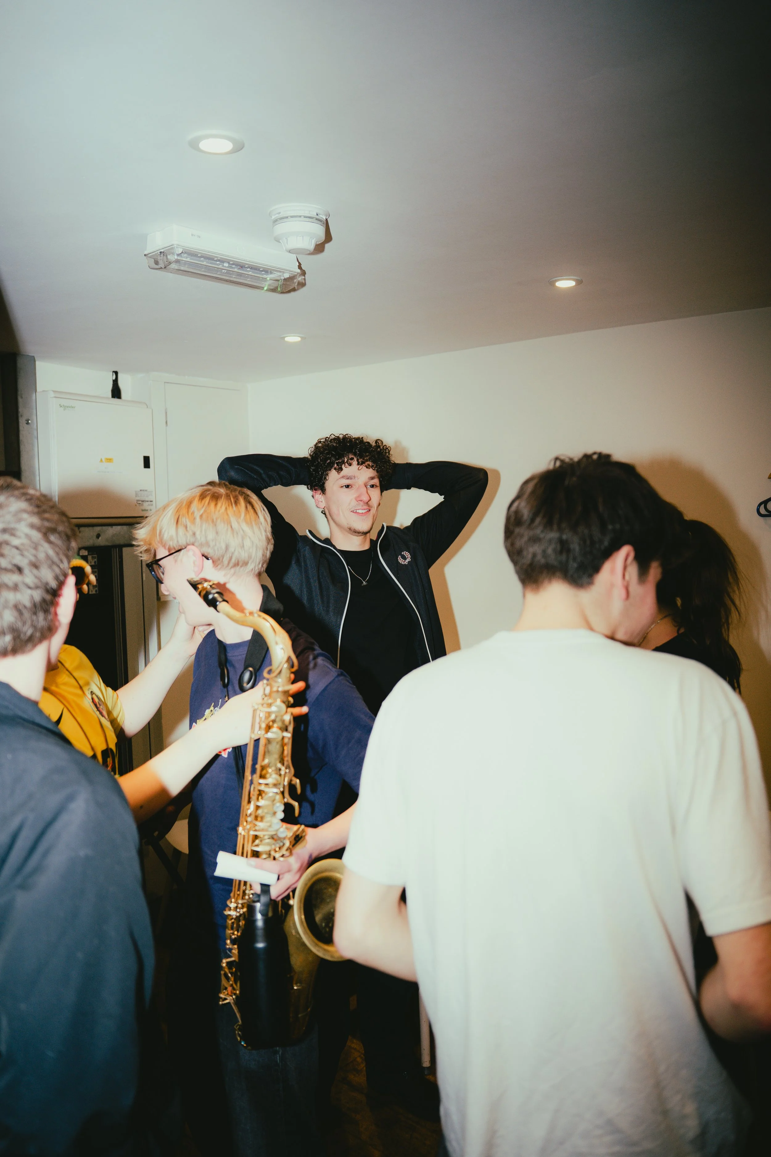 Group of young people socializing, one with sunglasses holding a saxophone, while others chat in a room with white walls and ceiling lights.