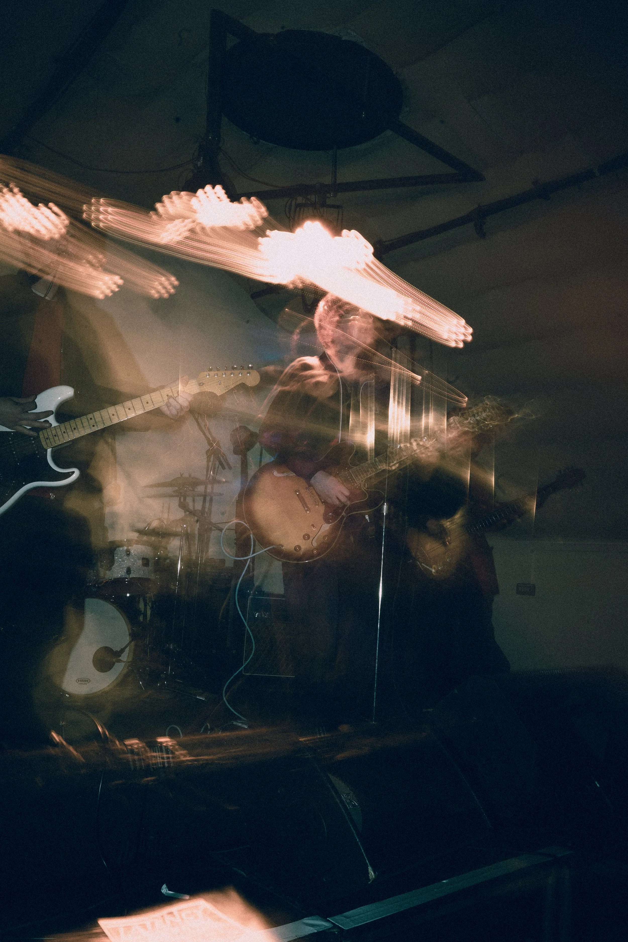 A blurry photo of a band performing on stage with guitars, drums, and colorful stage lights.