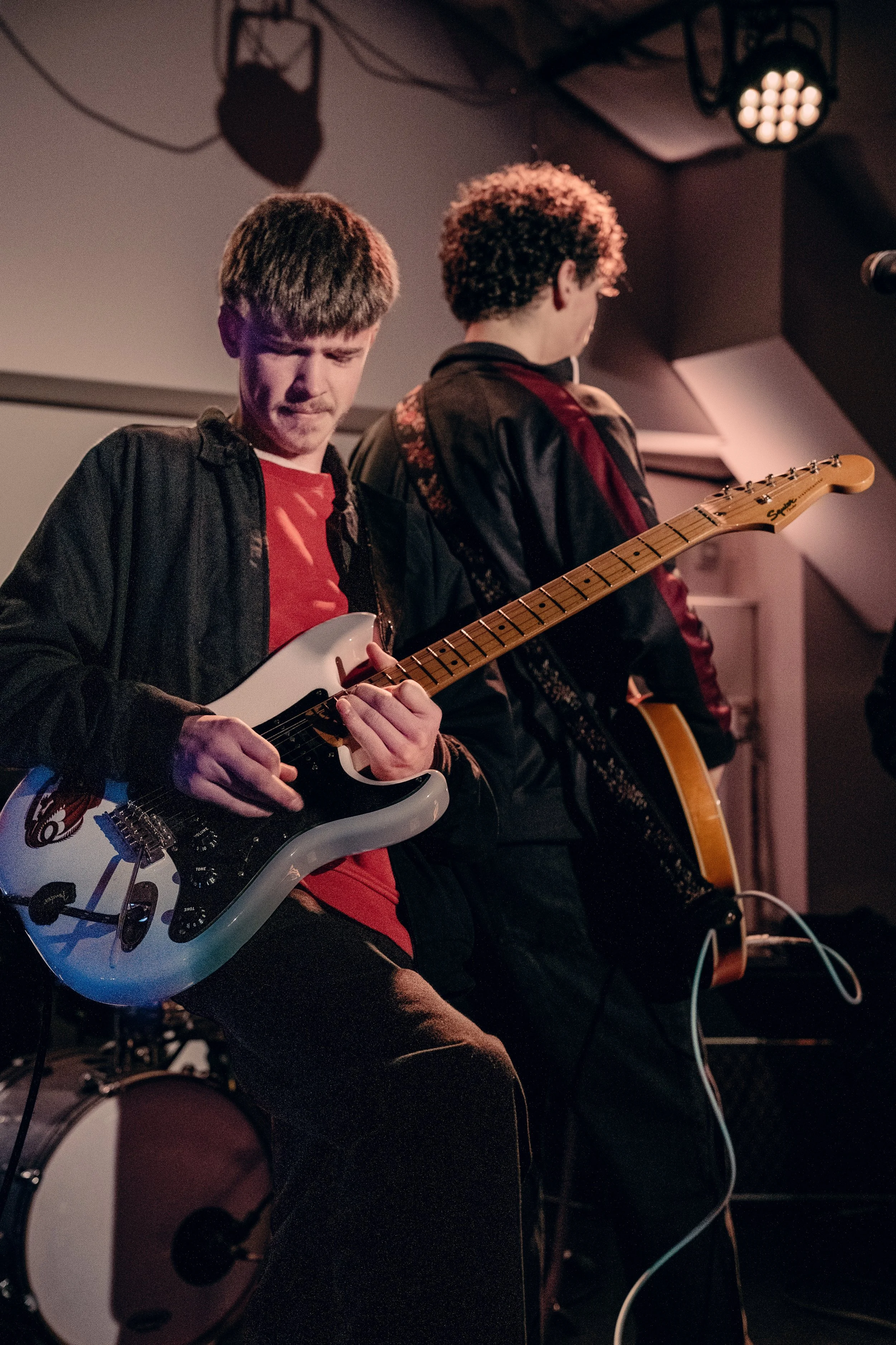 Two young men playing guitars on stage during a performance, one with an electric guitar and the other with an acoustic guitar, in a dimly lit indoor venue.
