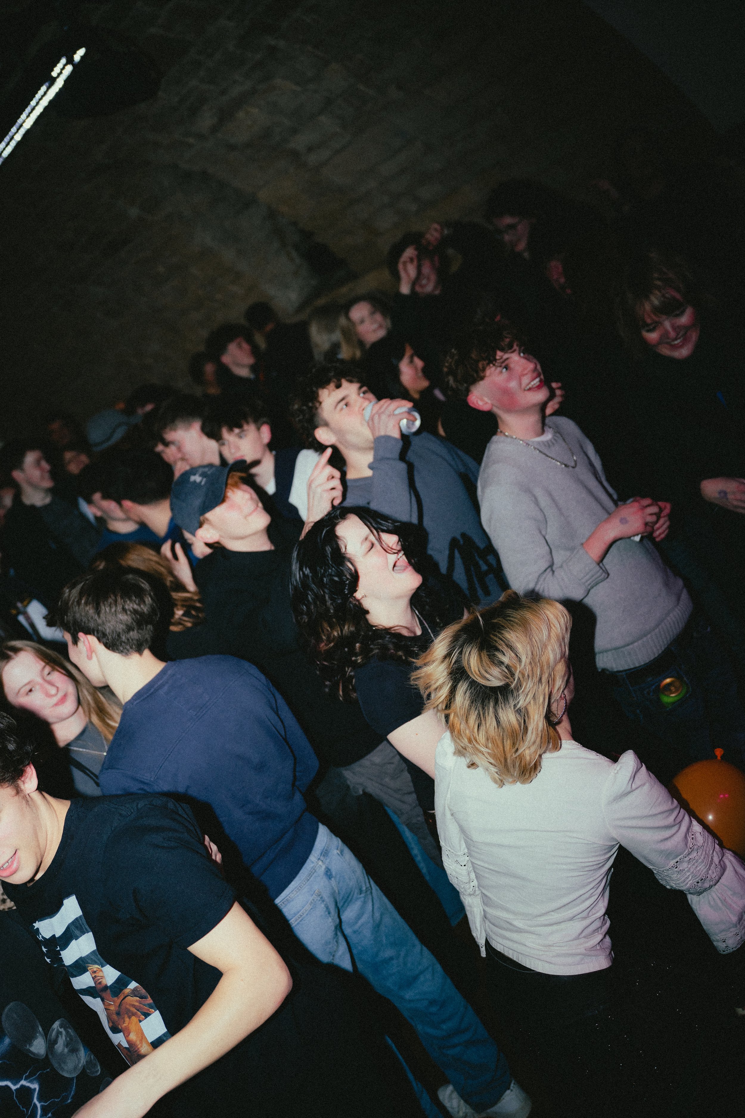 Group of young people enjoying a concert or party under a brick arch ceiling, some smiling, laughing, and dancing.