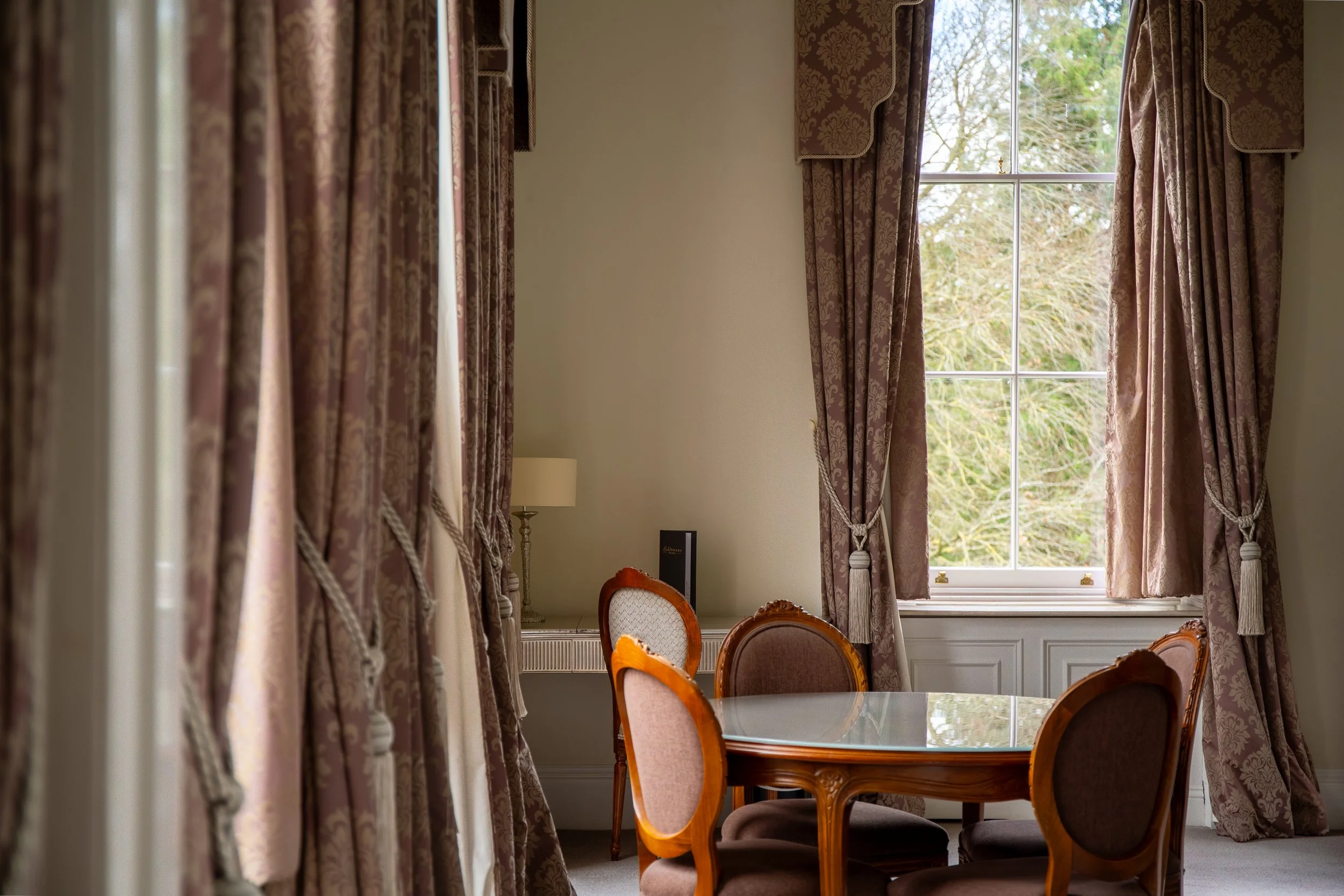 A dining room with a large window, elegant brown patterned curtains with tiebacks, a round wooden table with a glass top, and four upholstered chairs with curved wooden frames.