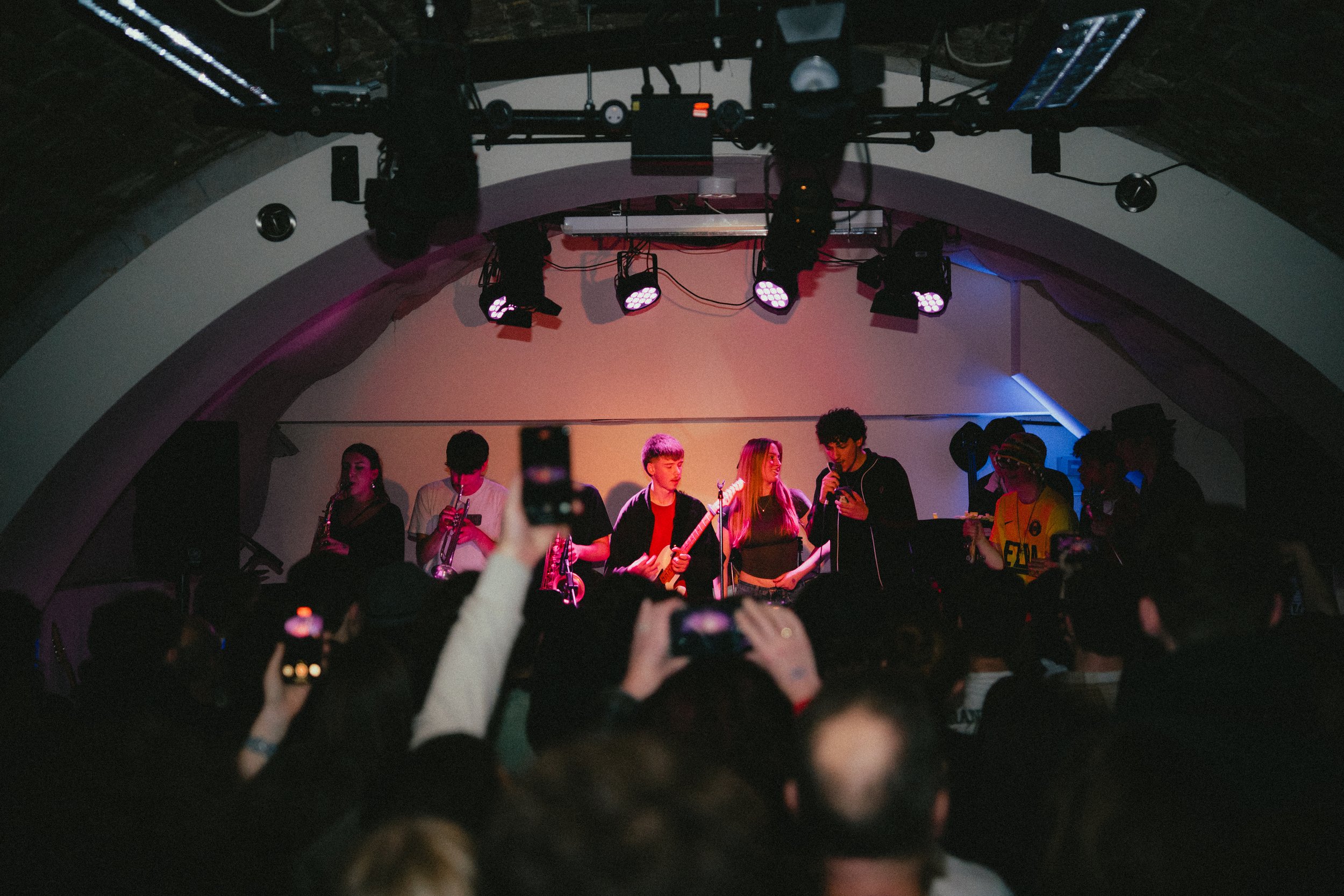 A live music performance on stage with a band, audience taking photos, and stage lighting in a dark venue.
