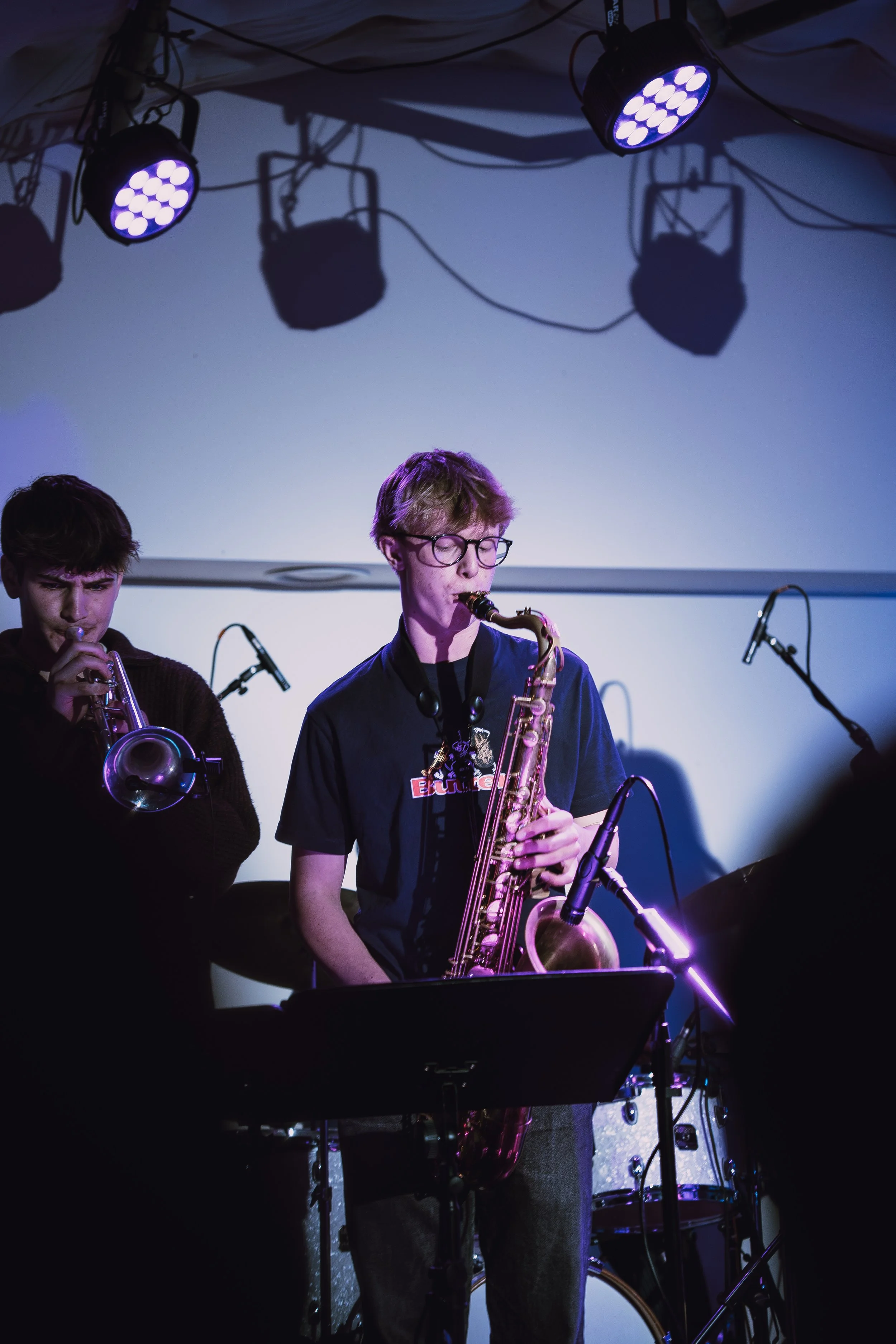 Young man playing a saxophone on stage with a microphone and a drum set in the background, stage lights overhead.