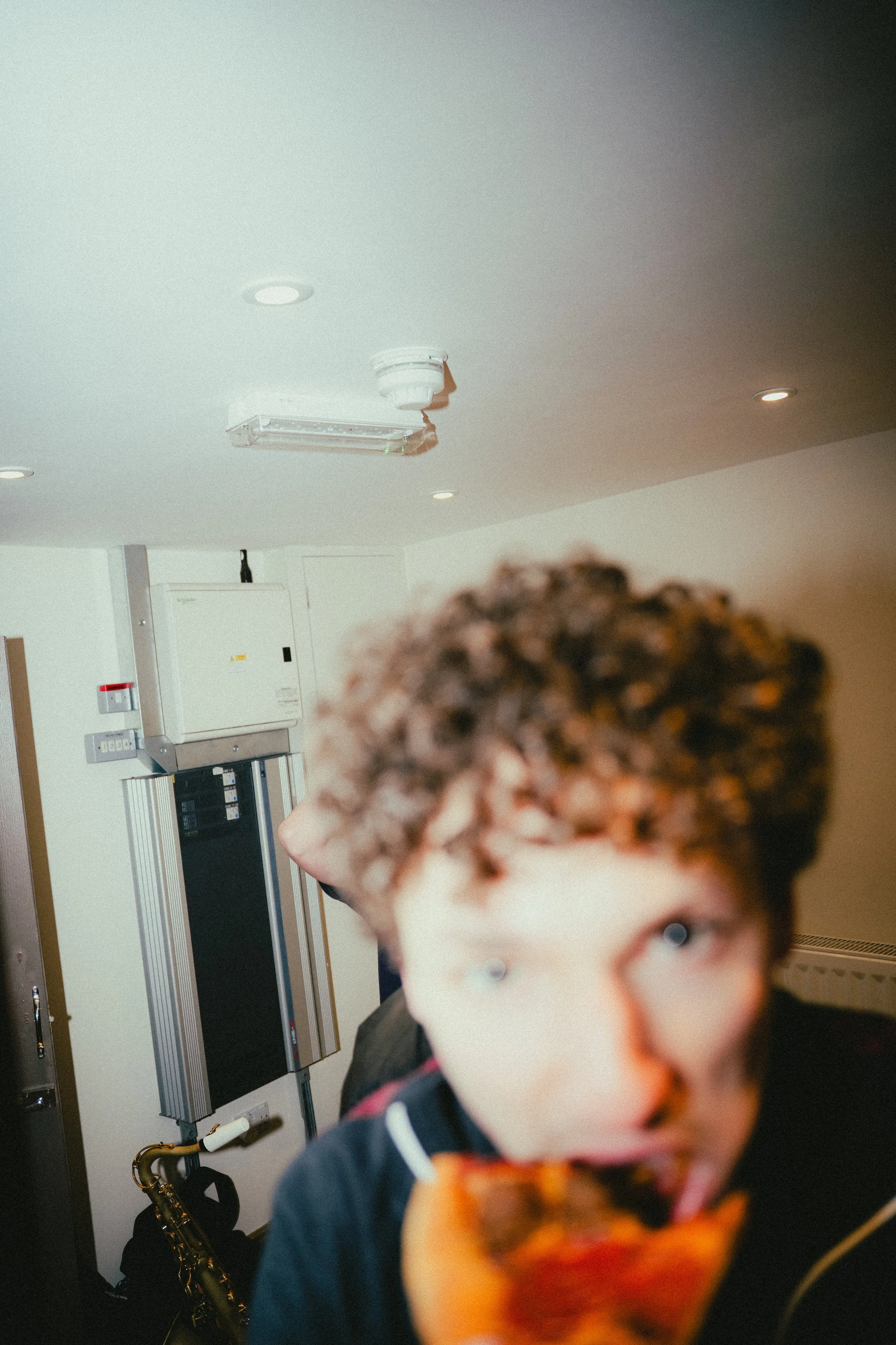 A young person with curly hair eating pizza in a room with a white ceiling and some electrical equipment on the wall.