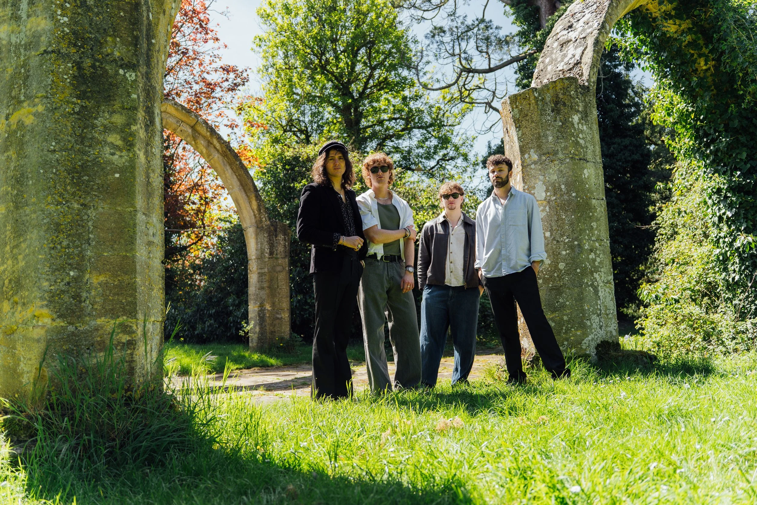 Four young adults standing outdoors near stone ruins in a lush, green park, with trees and a clear sky in the background.
