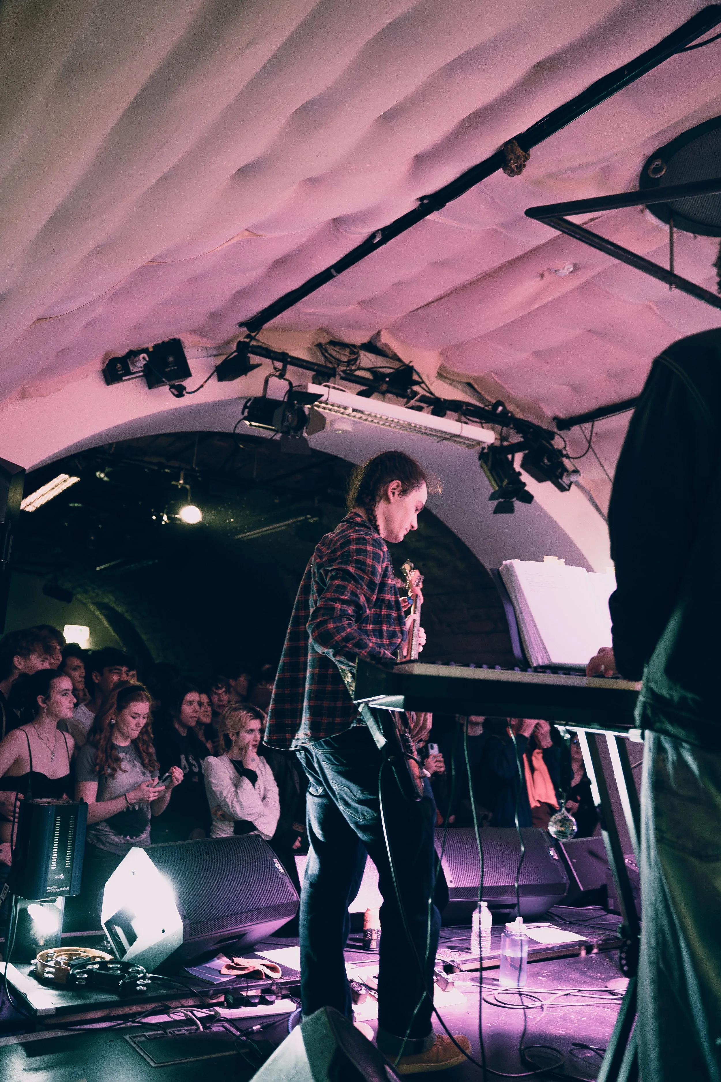A musician with braided hair playing a keyboard on stage during a concert, with a crowd of young people watching in the background.