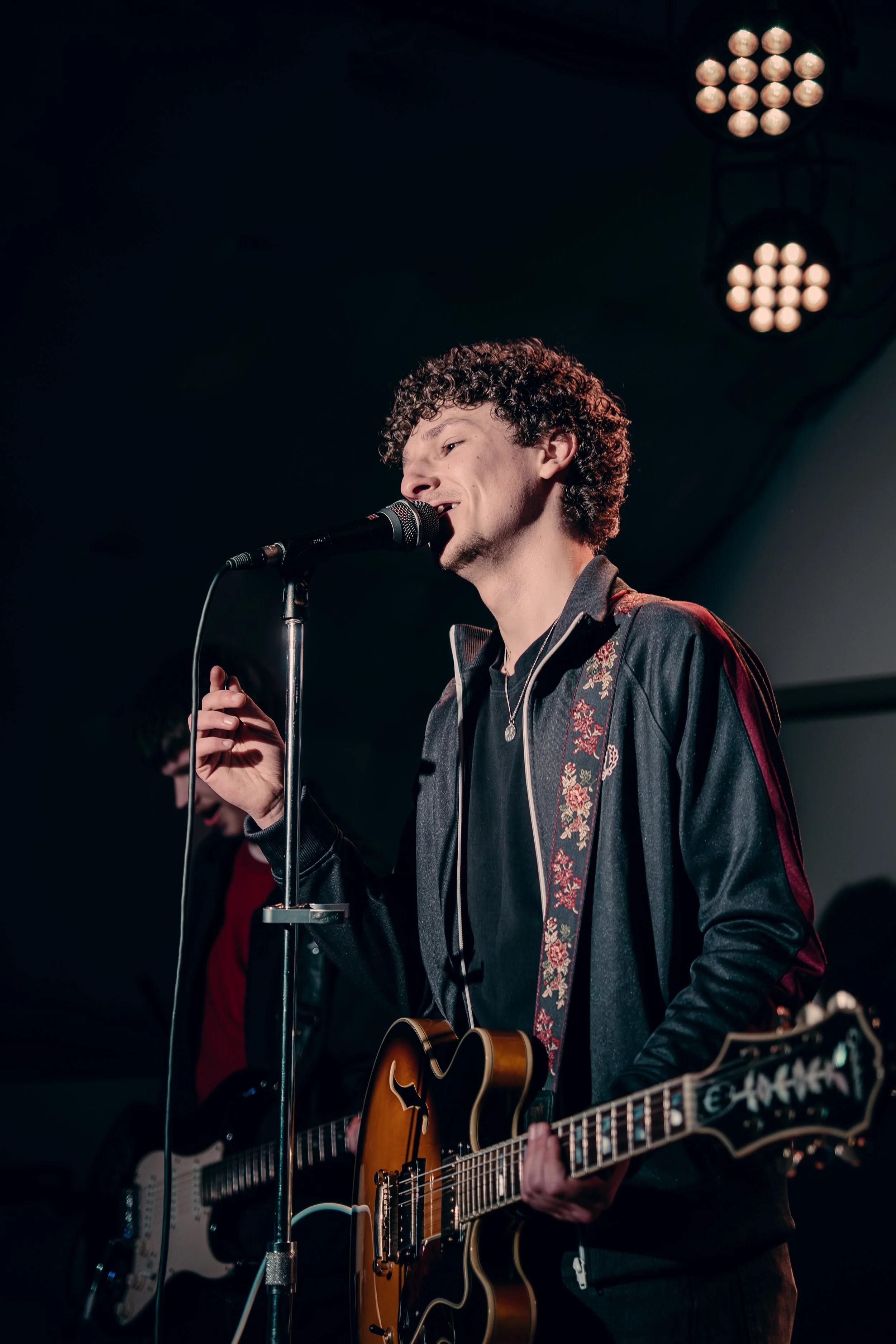 A young man with curly hair singing into a microphone while playing an electric guitar on stage, with stage lights overhead in the background.