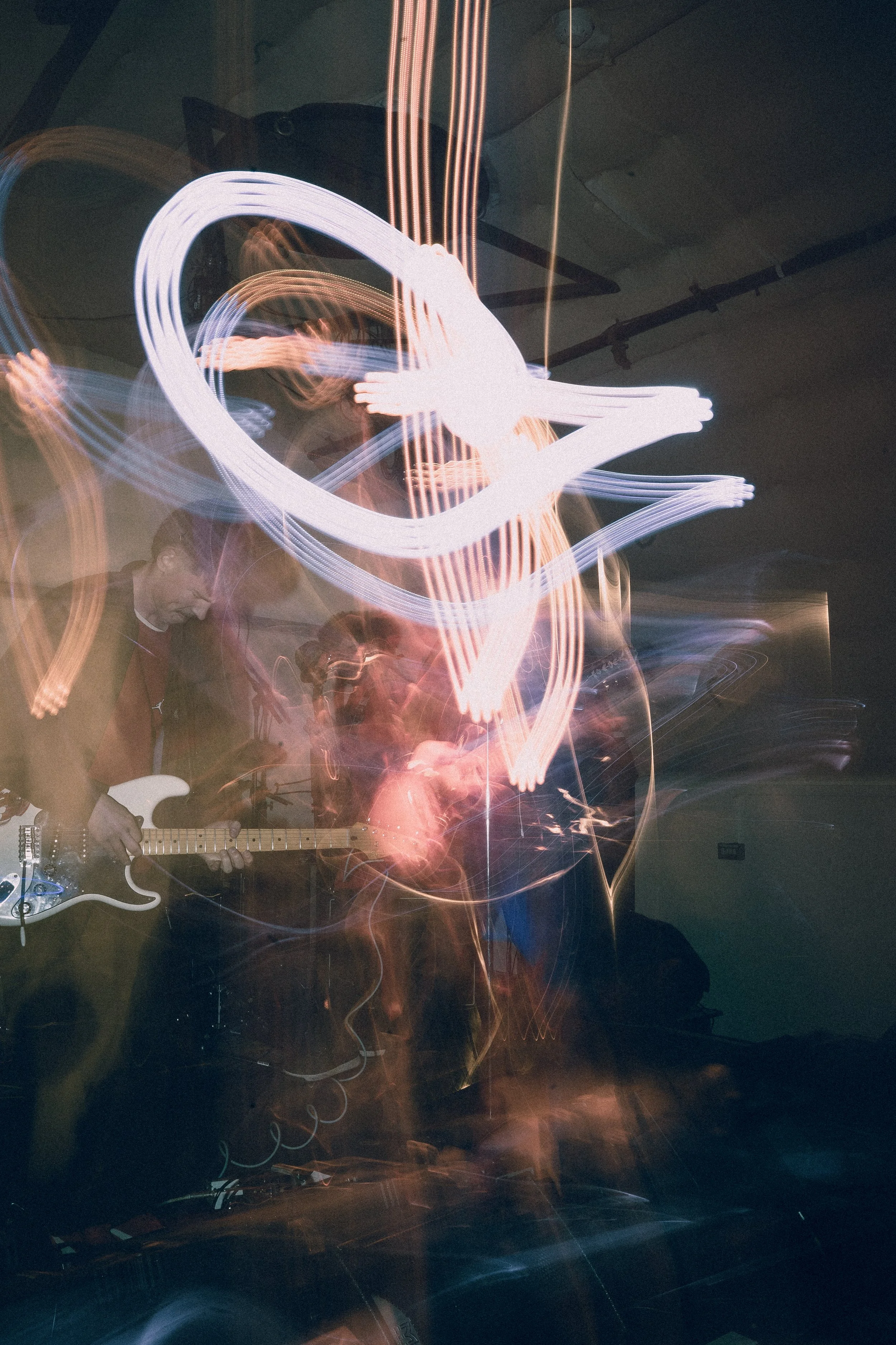 A long exposure photograph of a live music performance shows a person playing an electric guitar, surrounded by light trails and blurred figures, with colorful streaks and glowing lines in a dark room.