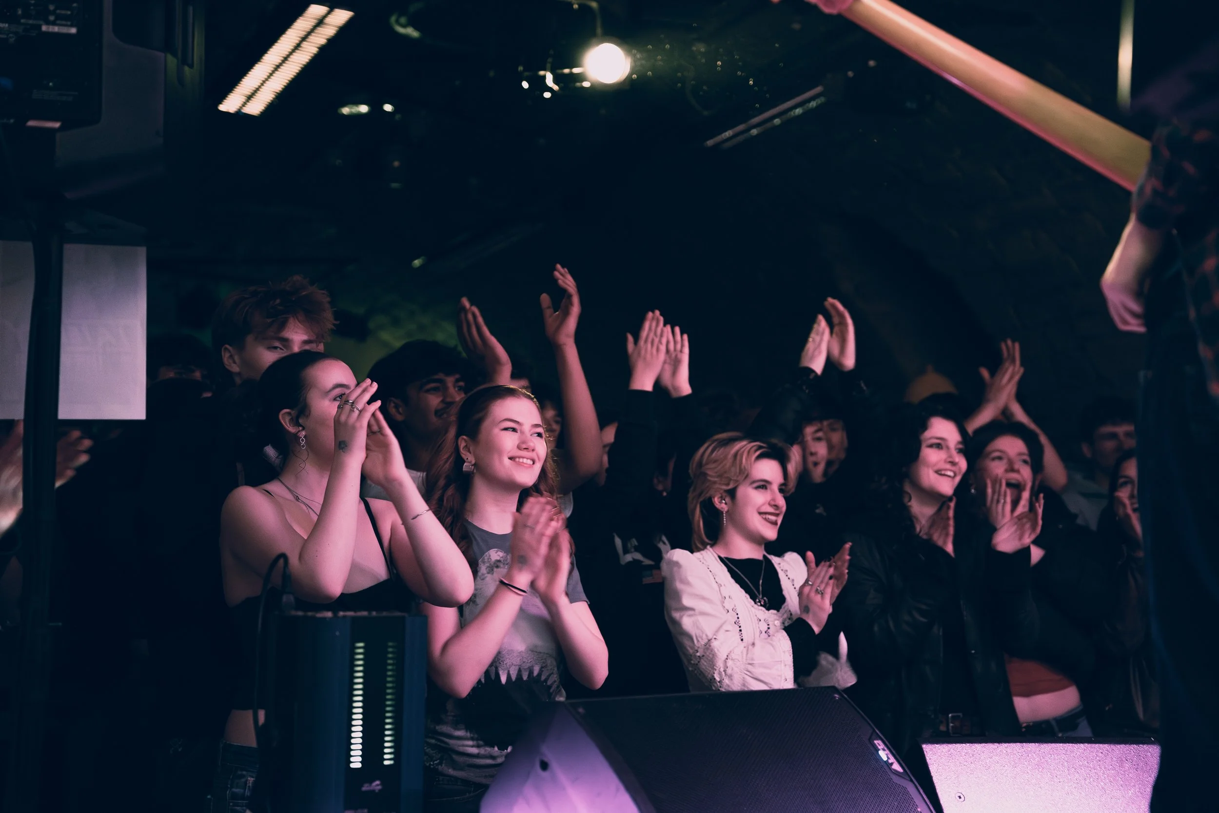 Audience at a concert or event clapping and smiling, with some raising their hands, inside a dimly lit venue.