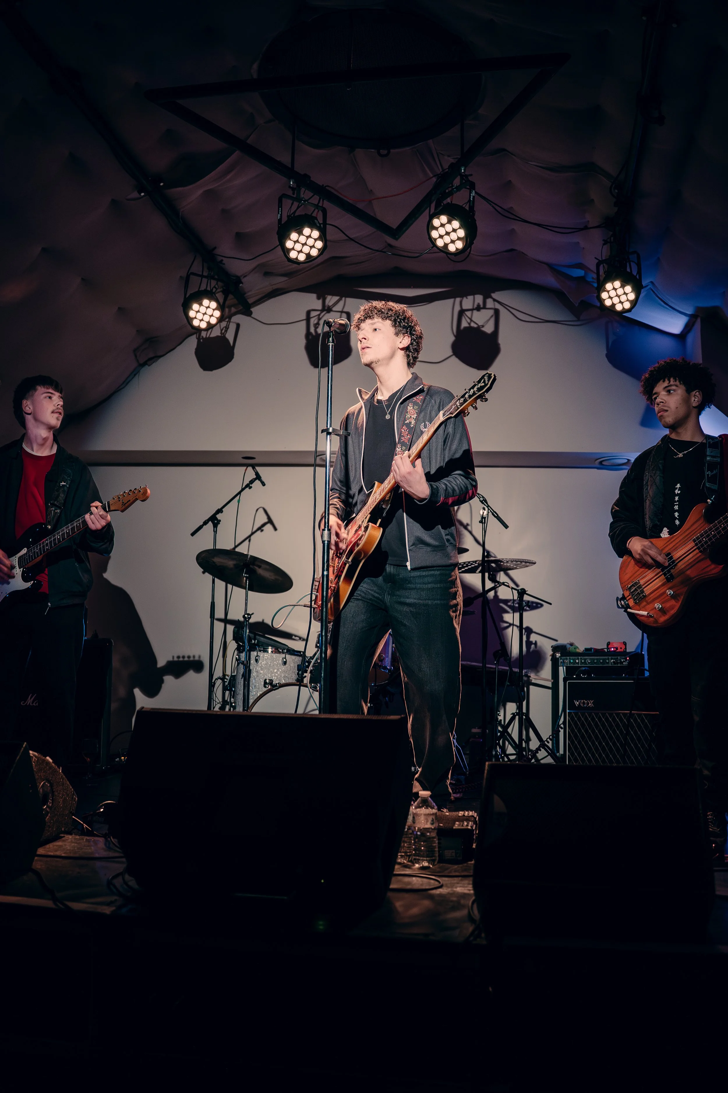 A band performing on stage with a lead singer playing guitar, two other guitarists, and drums in the background, under stage lights.
