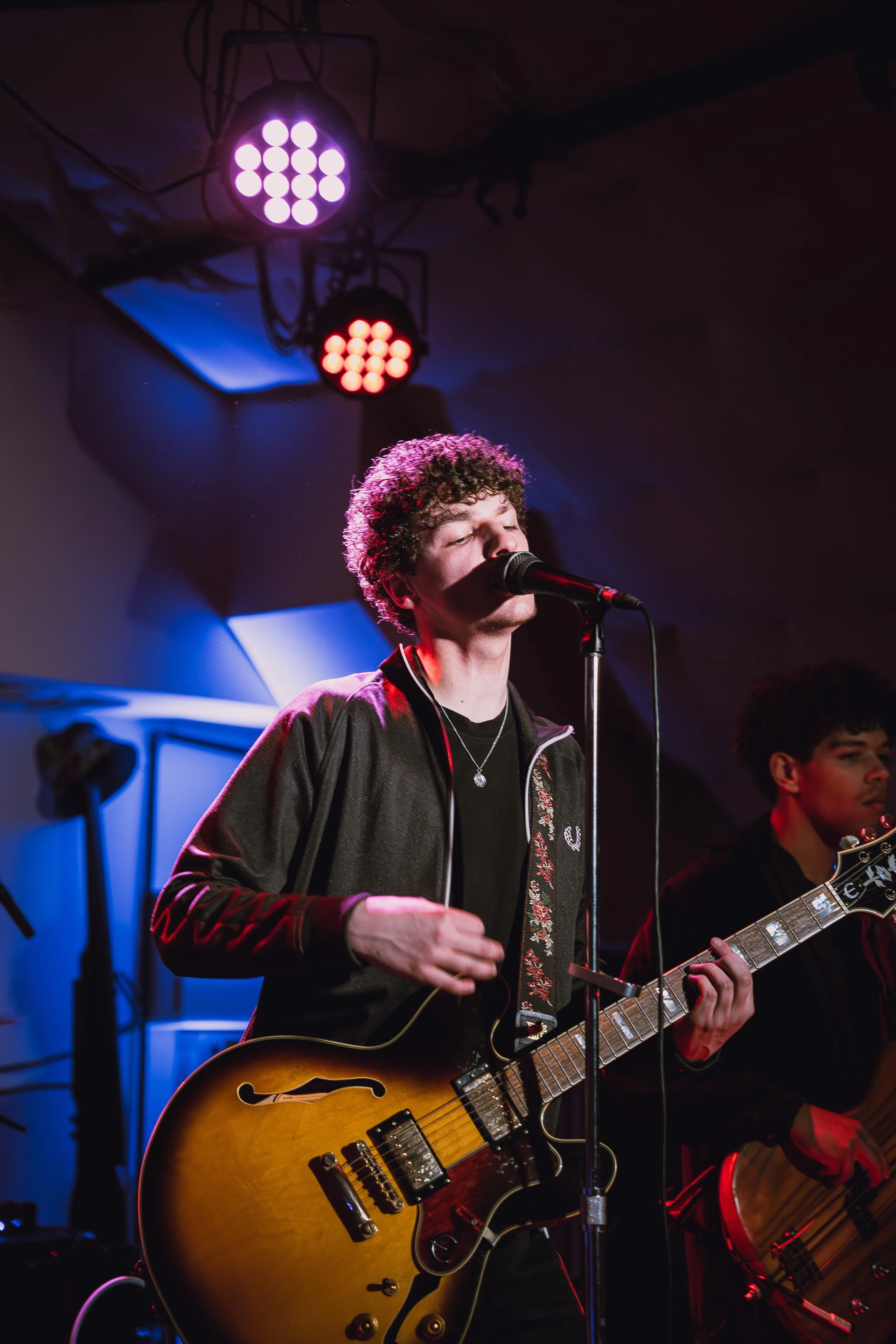 Young male musician singing into a microphone and playing an electric guitar on stage with colorful lights overhead.