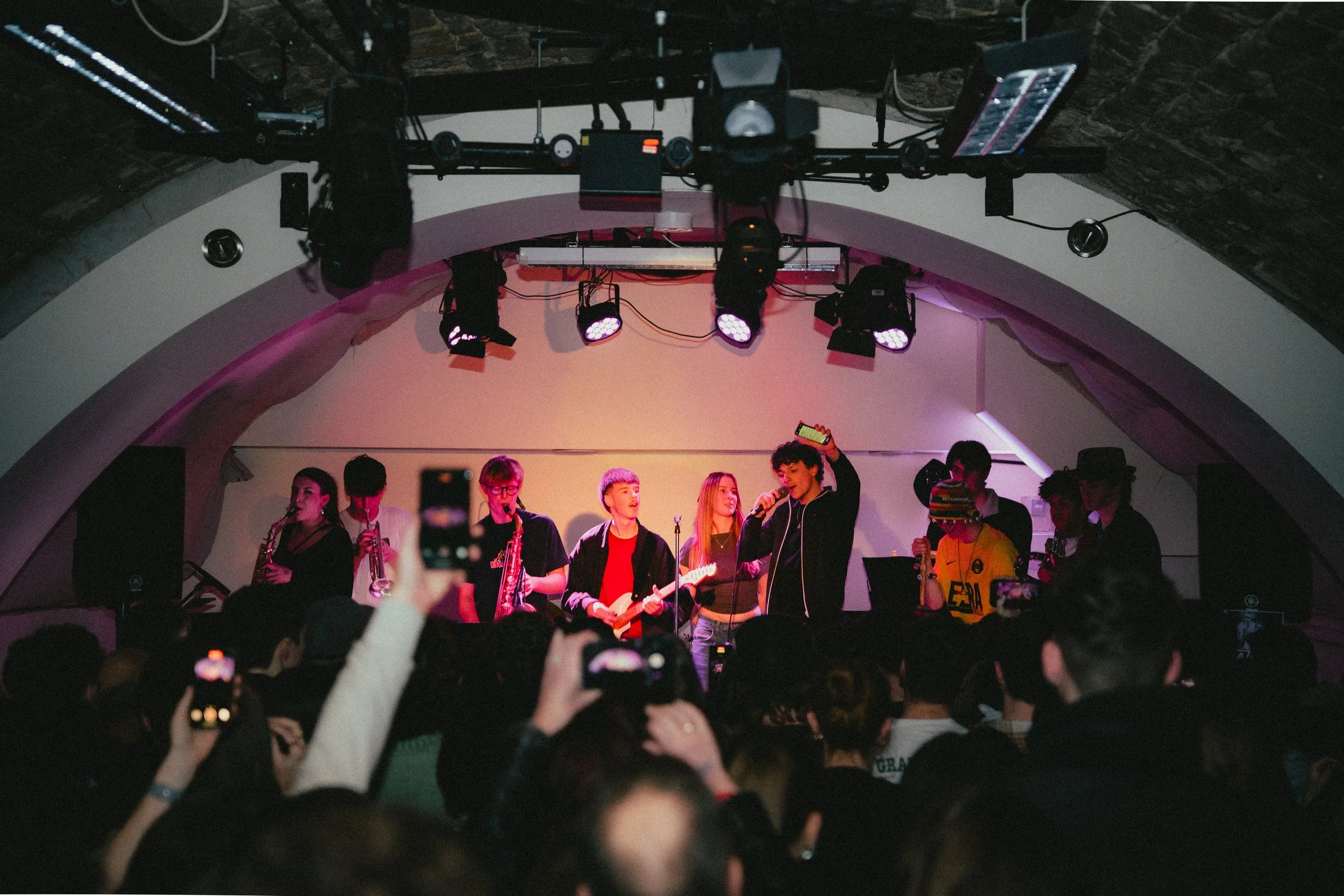 A band of musicians performing on stage in front of an audience, with some audience members taking photos. The stage is lit with pink and purple lights and has a curved ceiling.