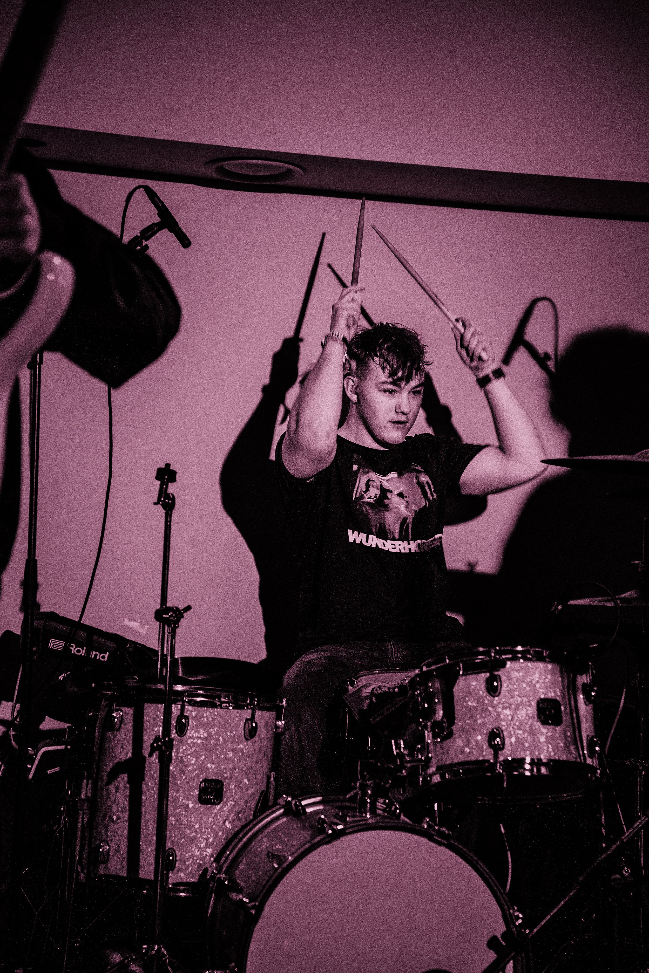 Young man playing drums on stage, wearing a black T-shirt with 'WUNDERHORSE' on it, holding drumsticks above his head, with recording equipment visible around him.