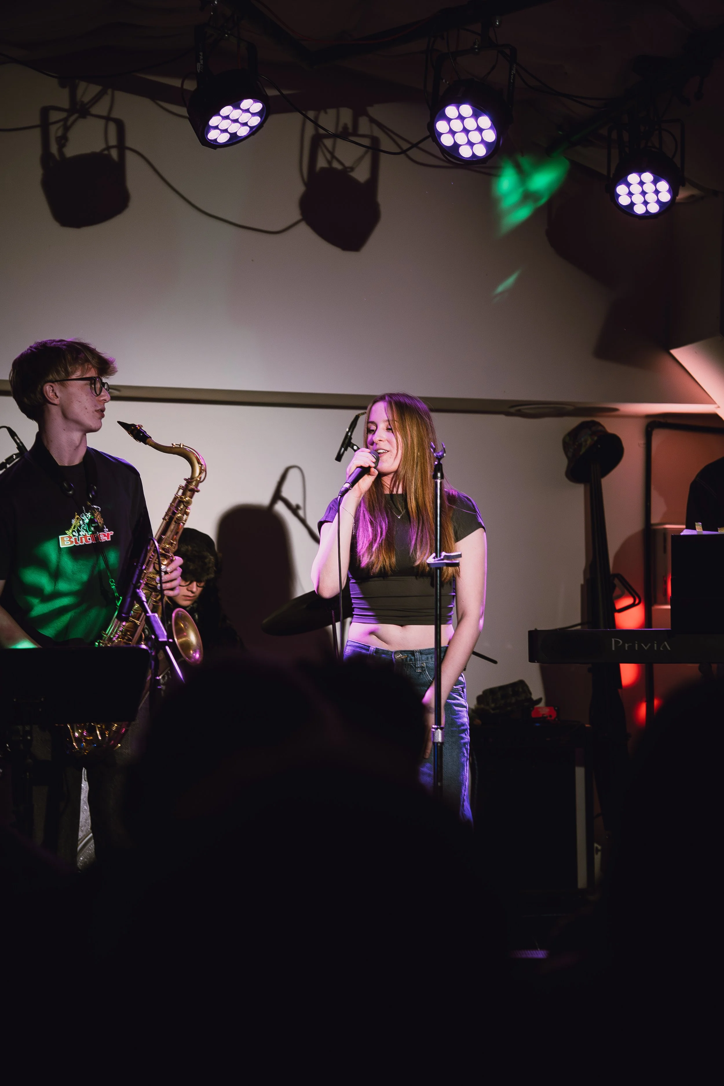 A young woman in a black crop top singing with a microphone on stage, accompanied by a saxophonist and other band members, under stage lights.