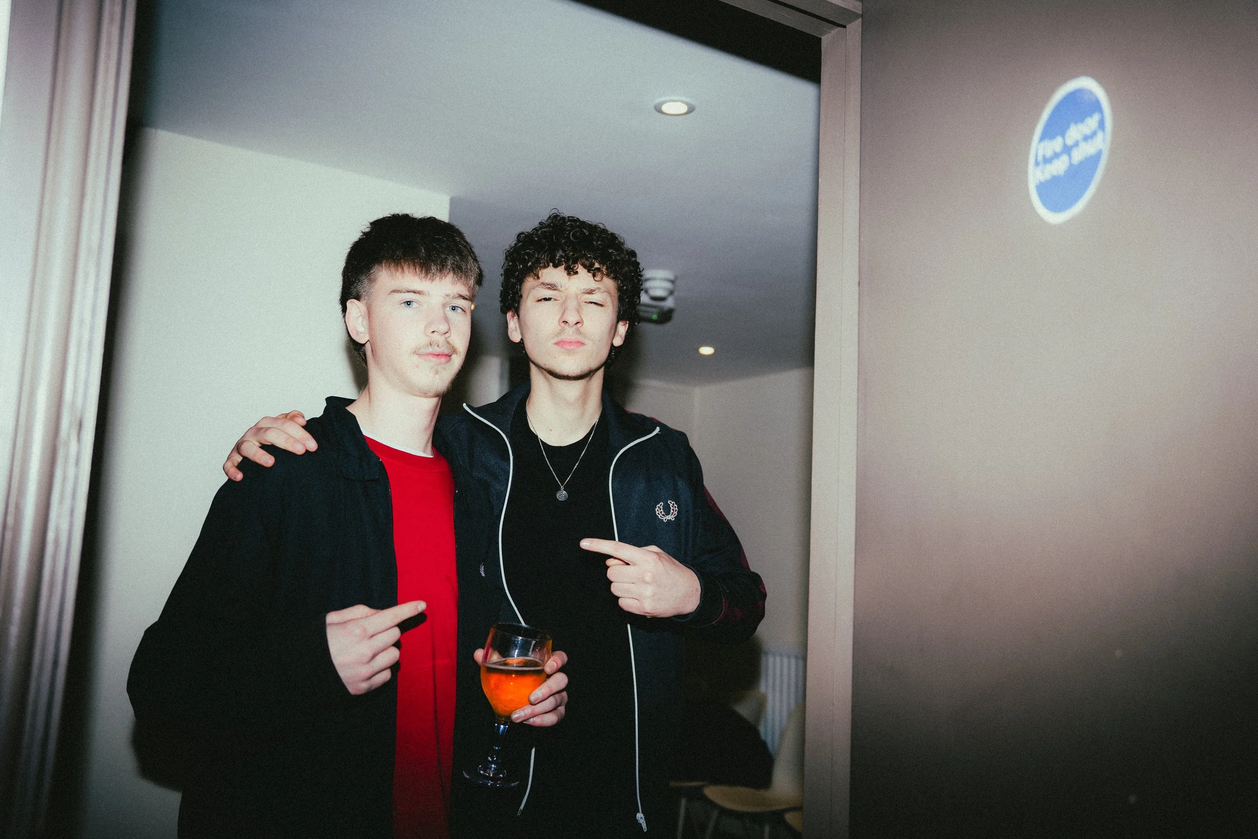 Two young men standing together, one holding a glass of orange-colored drink, both pointing at each other, inside a room with a doorway and a blue sign on the wall.