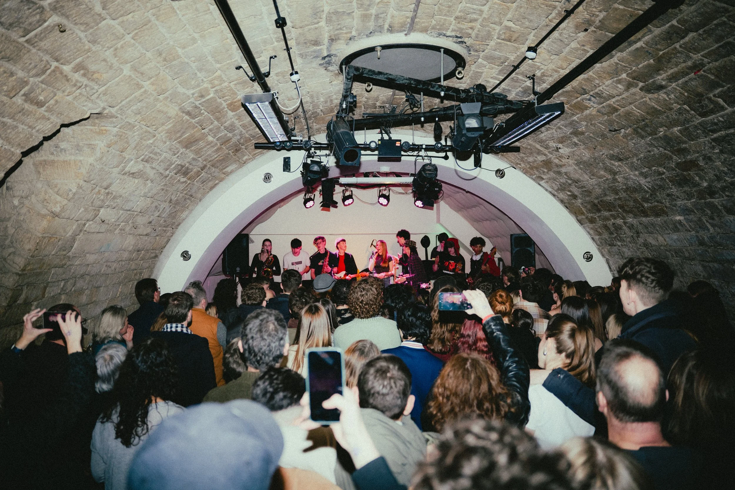 A live music performance in an underground venue with a stone arch ceiling. Musicians are on stage with instruments, and the audience is watching, some recording with their phones.