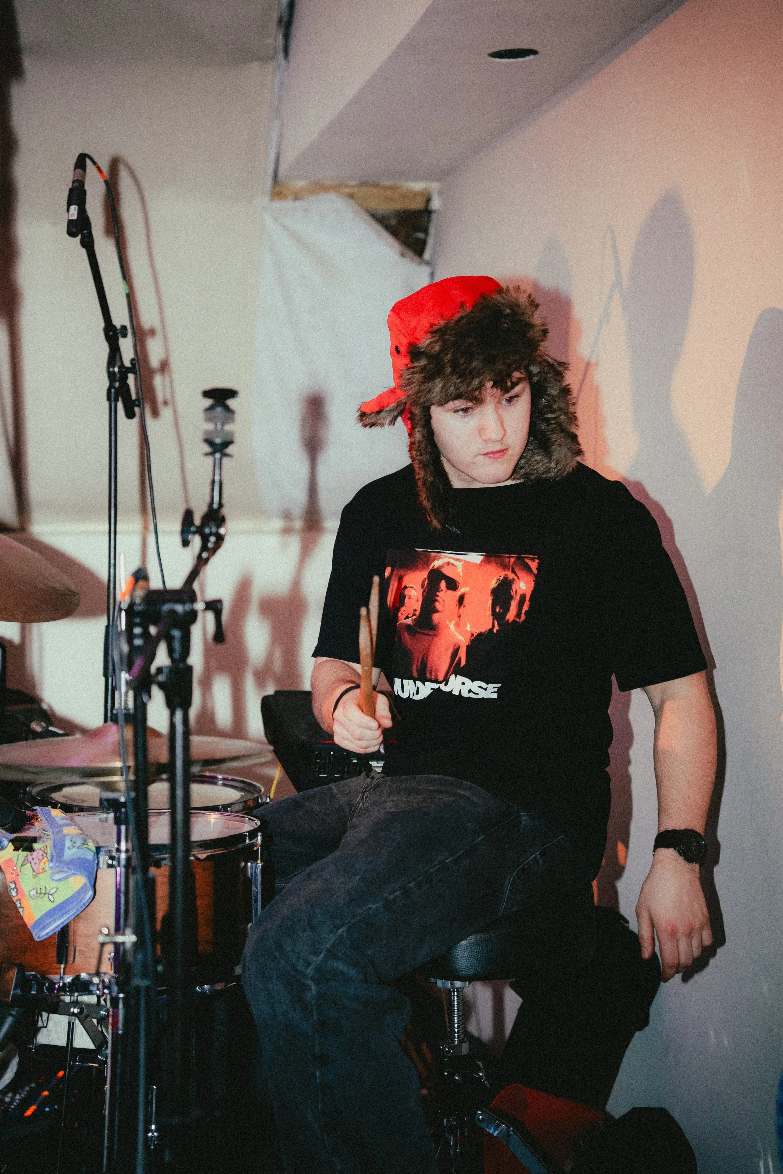 A young man wearing a red hat with ear flaps and a black T-shirt featuring band members, sitting on a stool with a drum set nearby, in a room with white and beige walls.