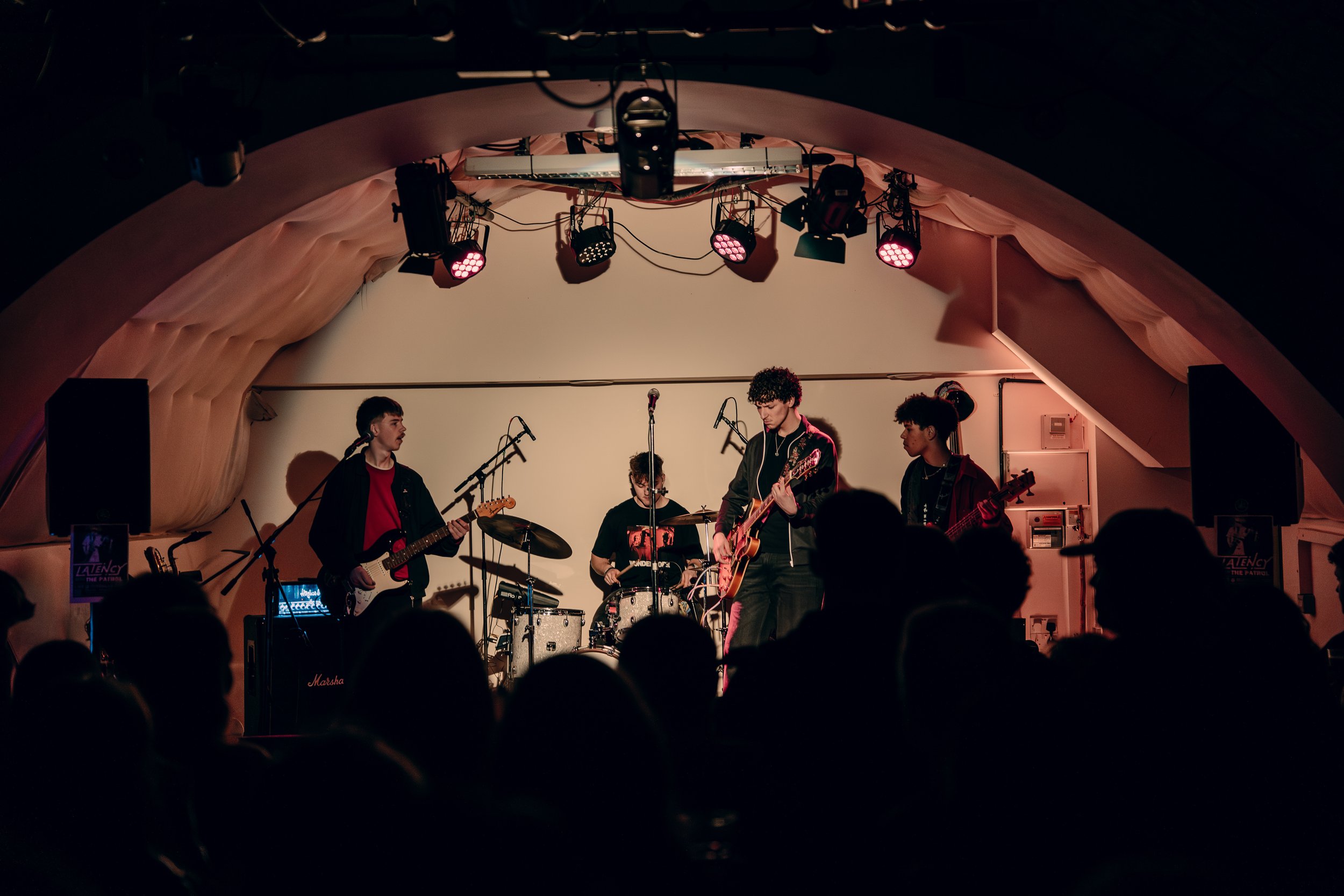 A band of four young men performing live on stage in a small venue, with one playing guitar, one on drums, another on bass, and a lead singer also playing guitar, under pink stage lighting.