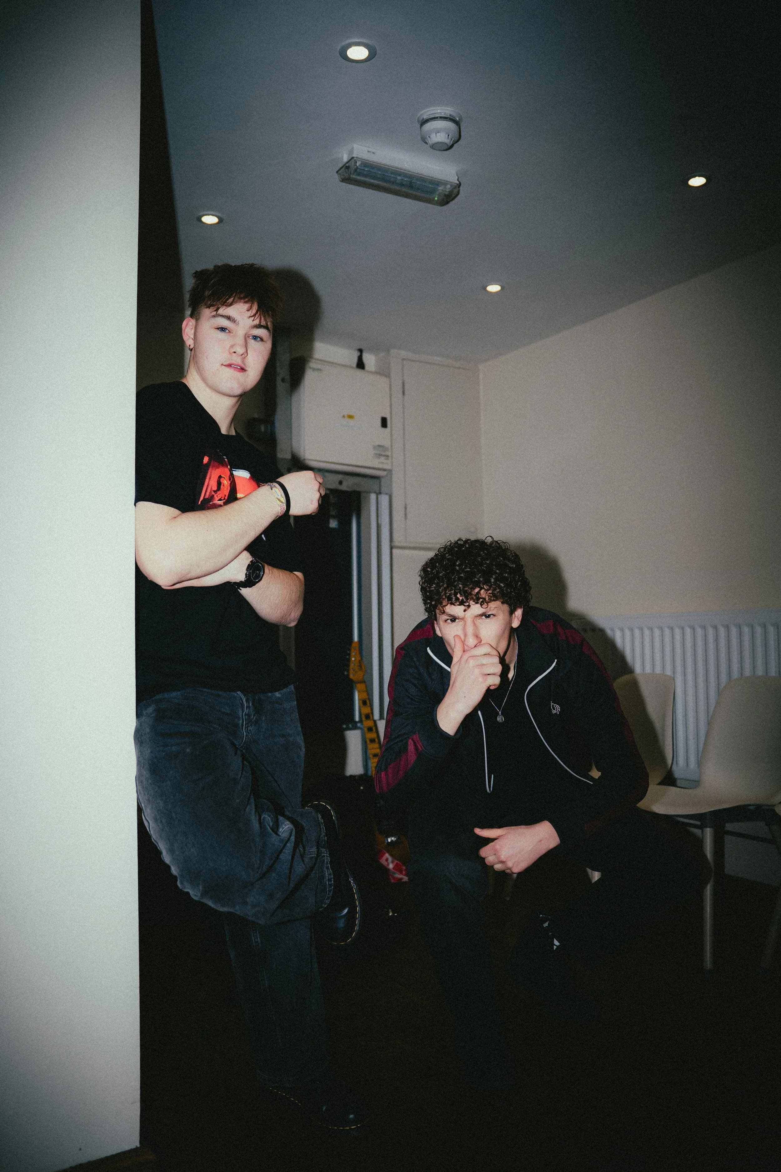 Two young men in a room with beige walls, one standing and one sitting, with guitar and chairs in the background, and ceiling lights.