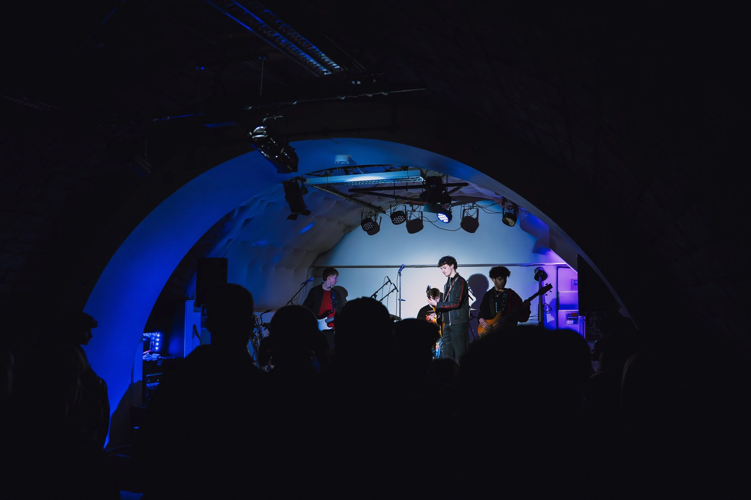 A band performs on stage in a dark venue, with purple and blue lighting, and an audience watching.