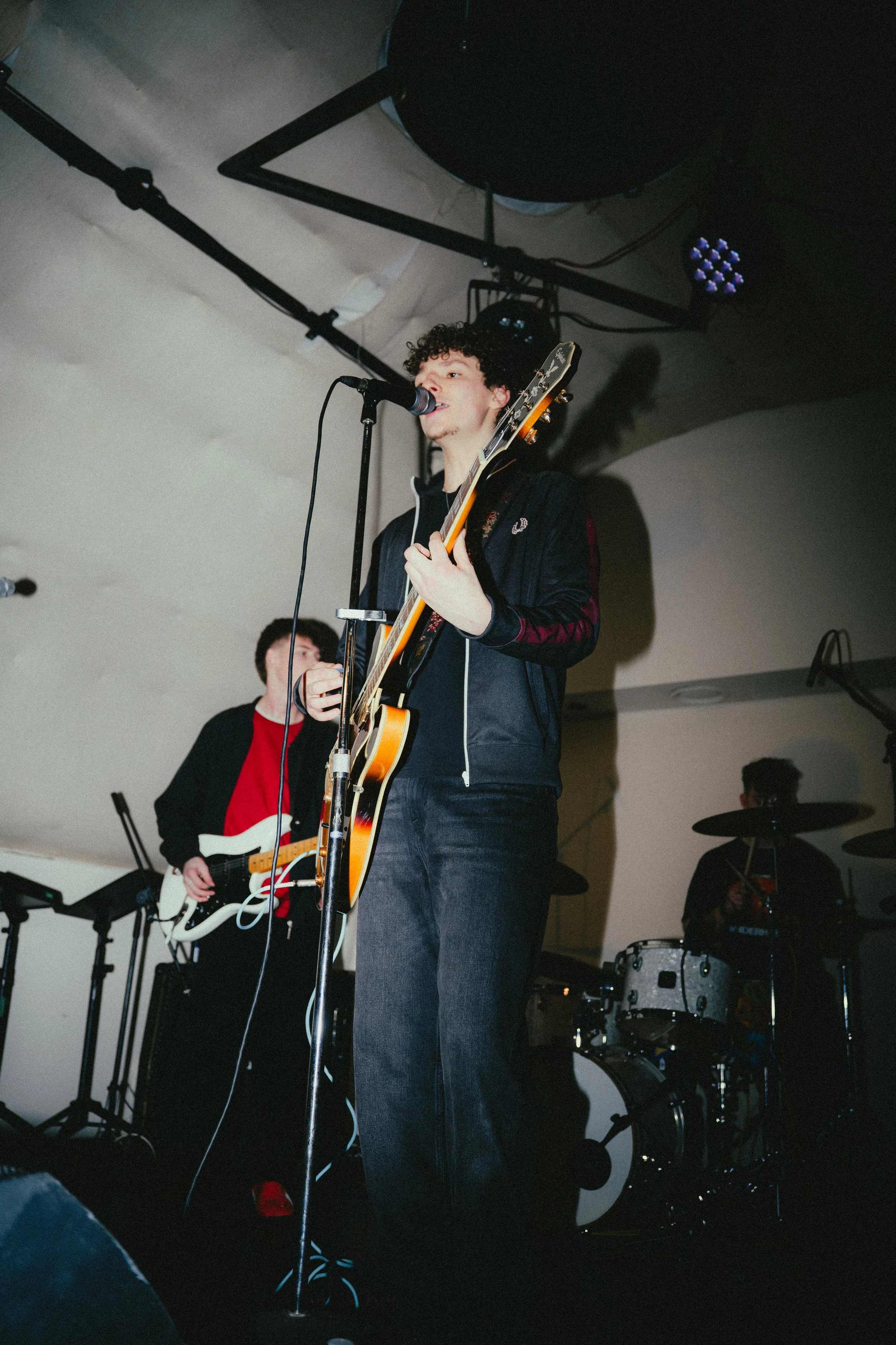 Young male musician with curly hair, singing into a microphone while playing an electric guitar on stage, with a band in the background.