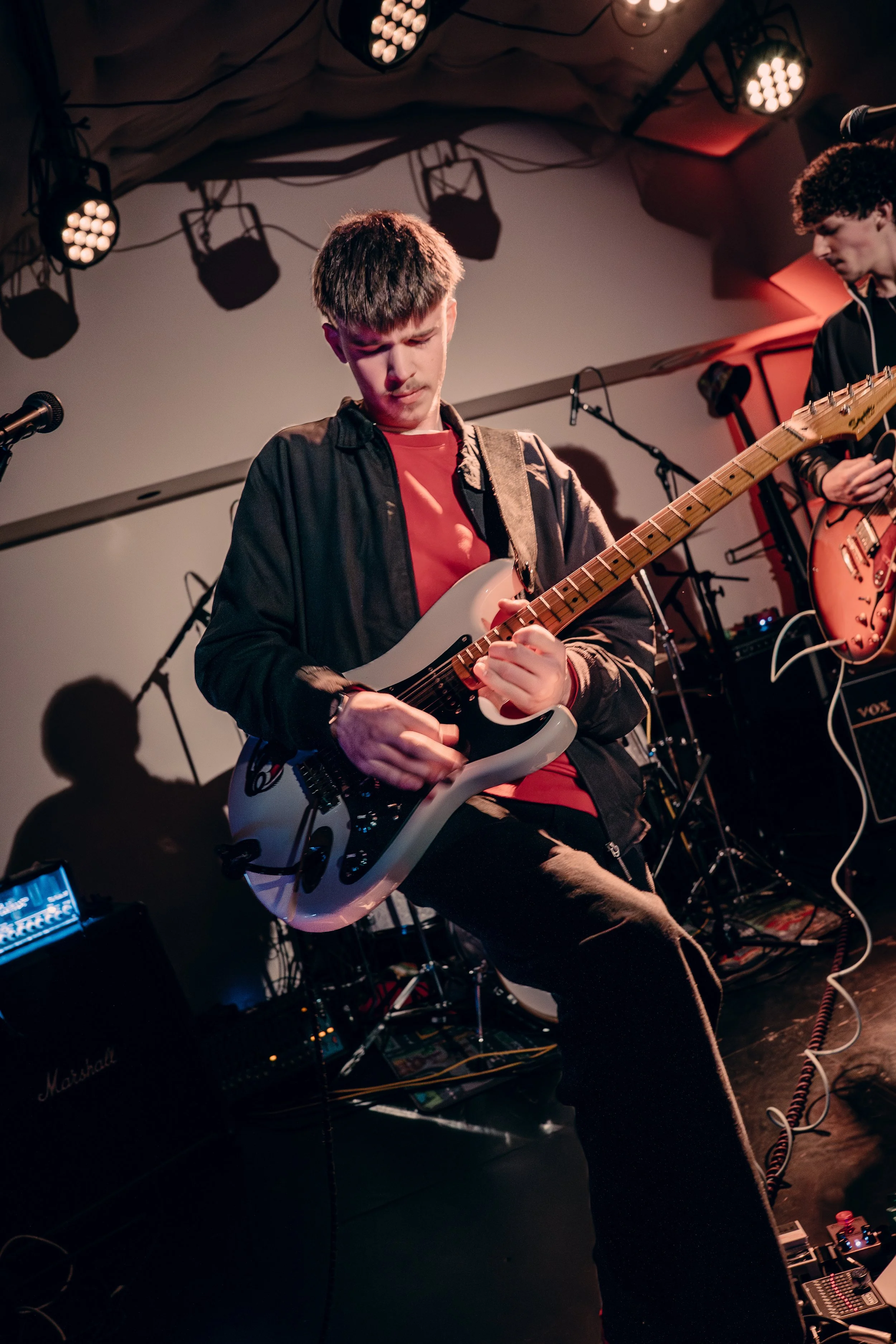 A young man playing an electric guitar on stage with stage lights overhead and musical equipment in the background.