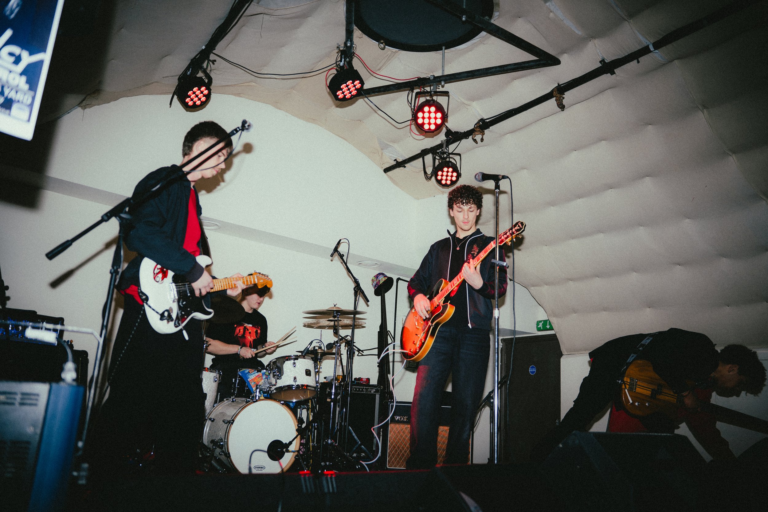 Four musicians on stage playing guitars and drums beneath red stage lights with a curved ceiling in a small venue.