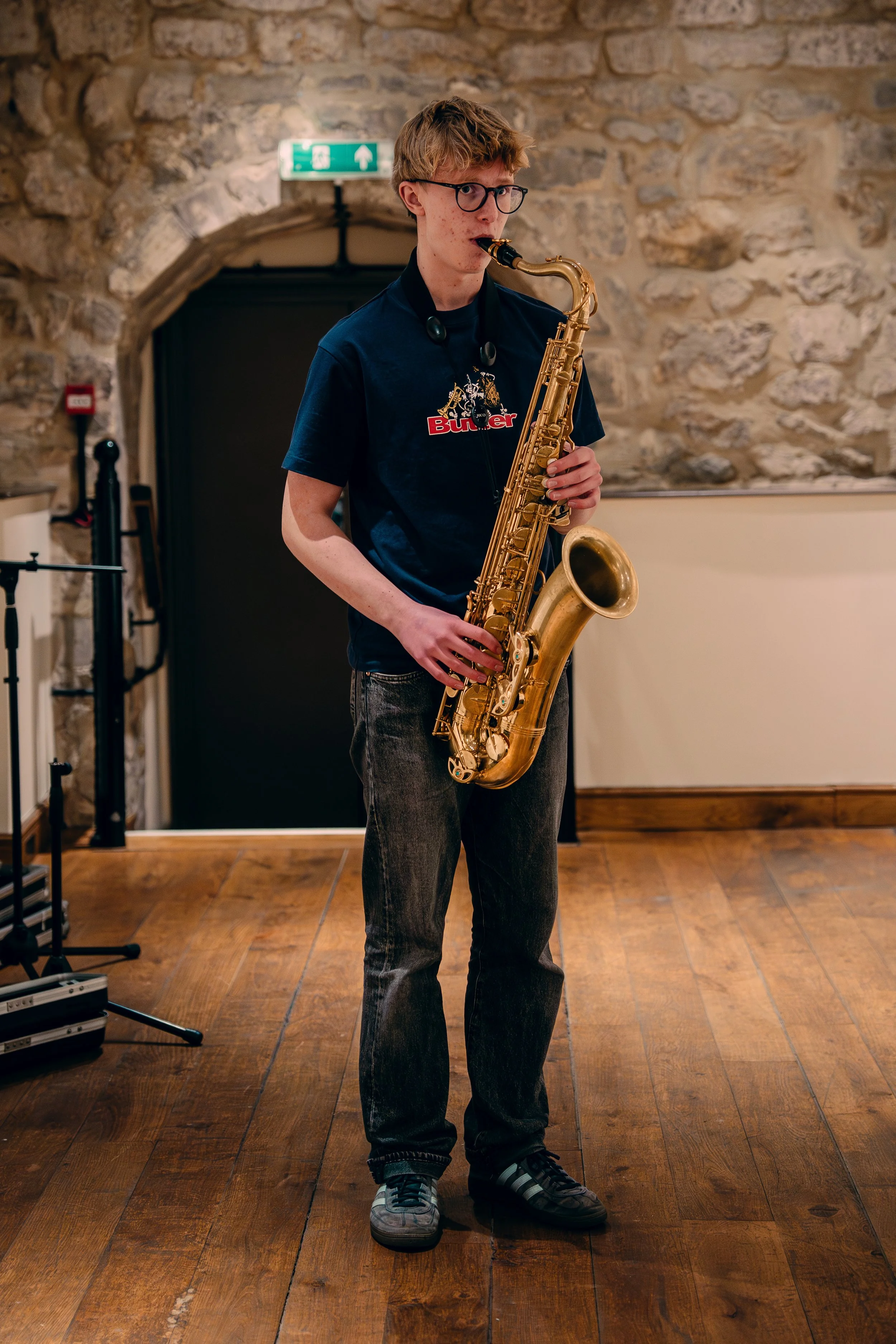 A young man with glasses and short blond hair playing a gold-colored saxophone indoors with a stone wall background.