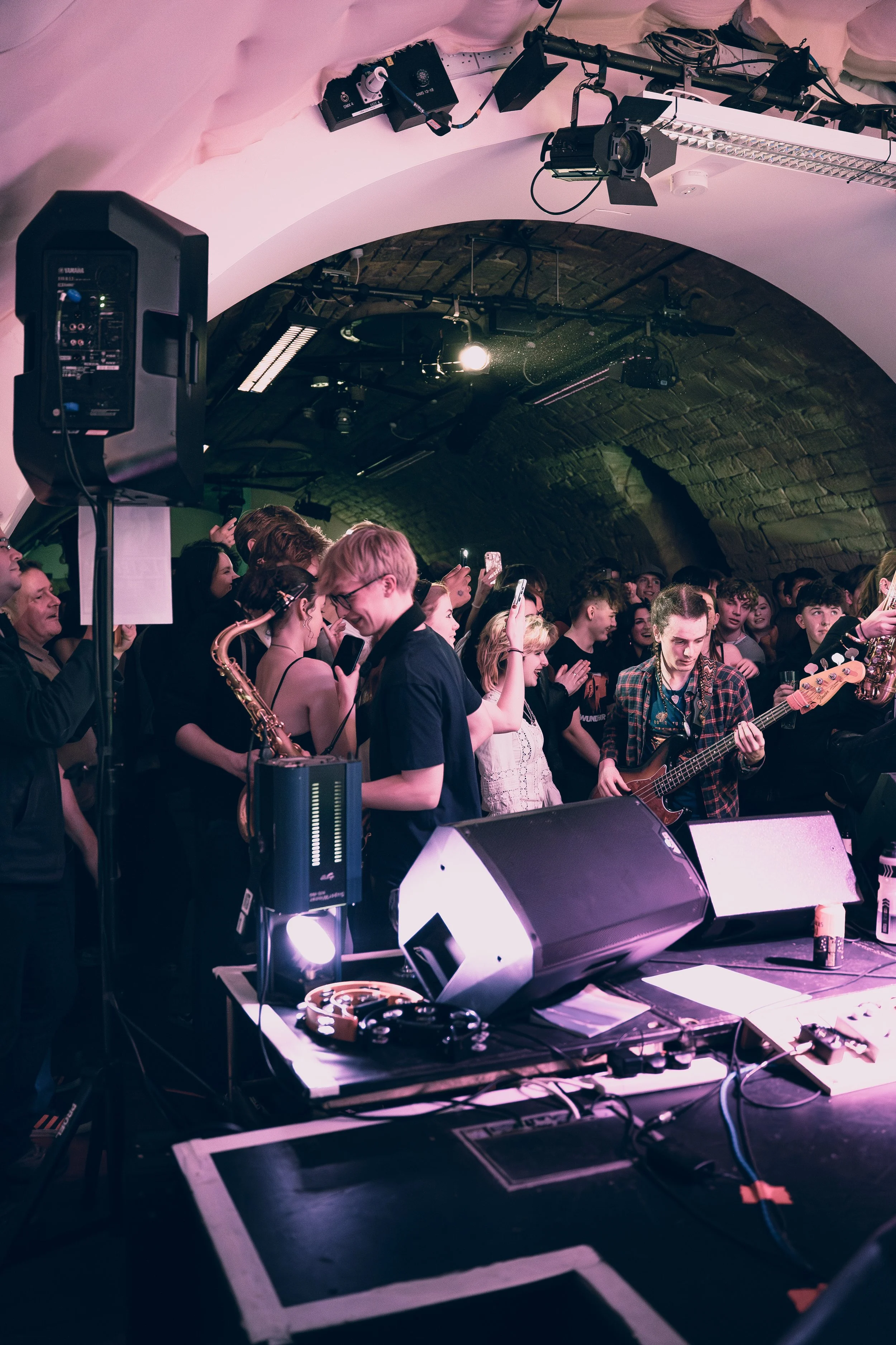 A crowd of people at a concert in a small underground venue with a brick arch ceiling, some holding up phones, with musical instruments and speakers on stage.