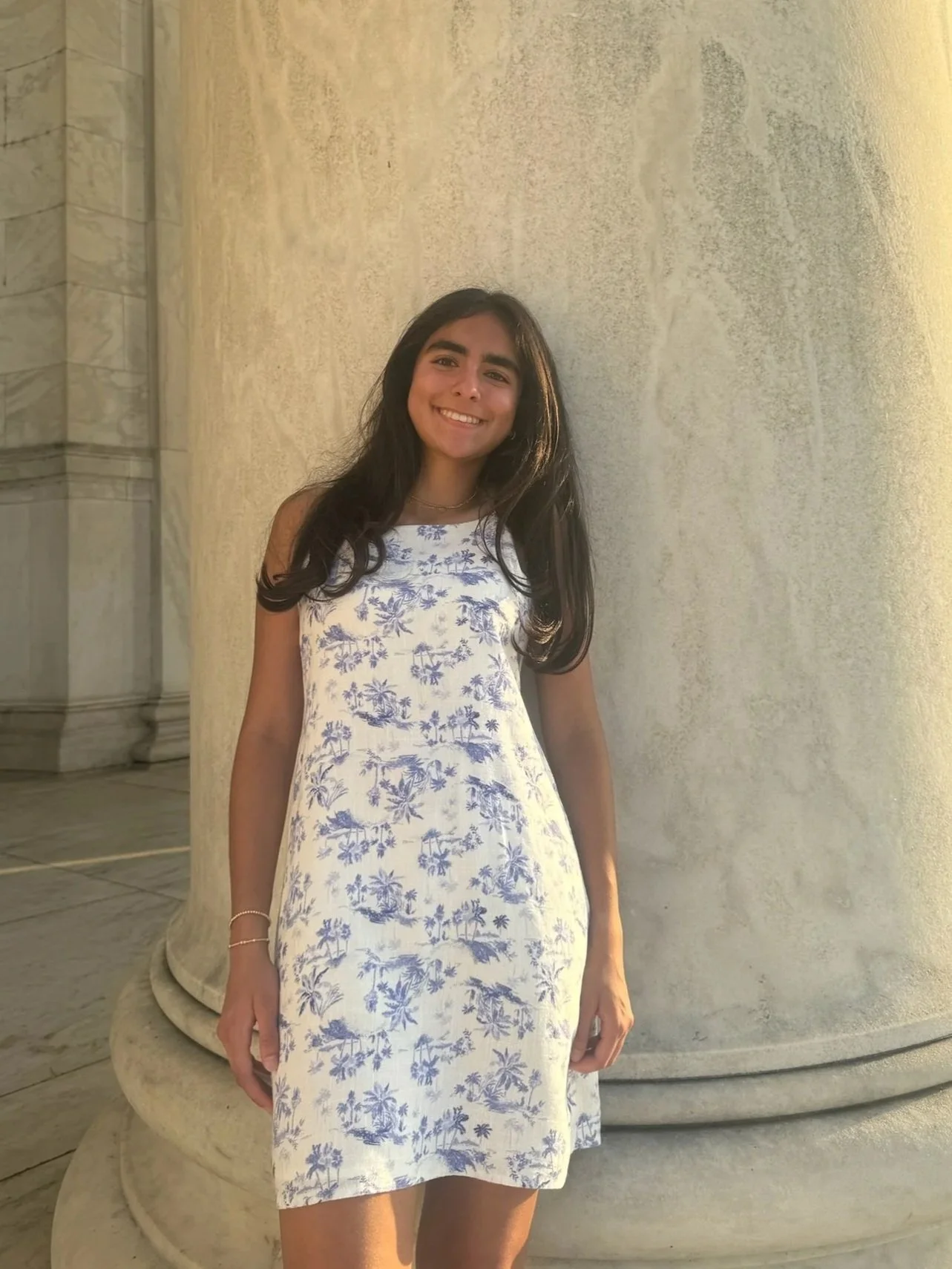 A young woman in a white and blue floral dress smiling and standing against a stone column.