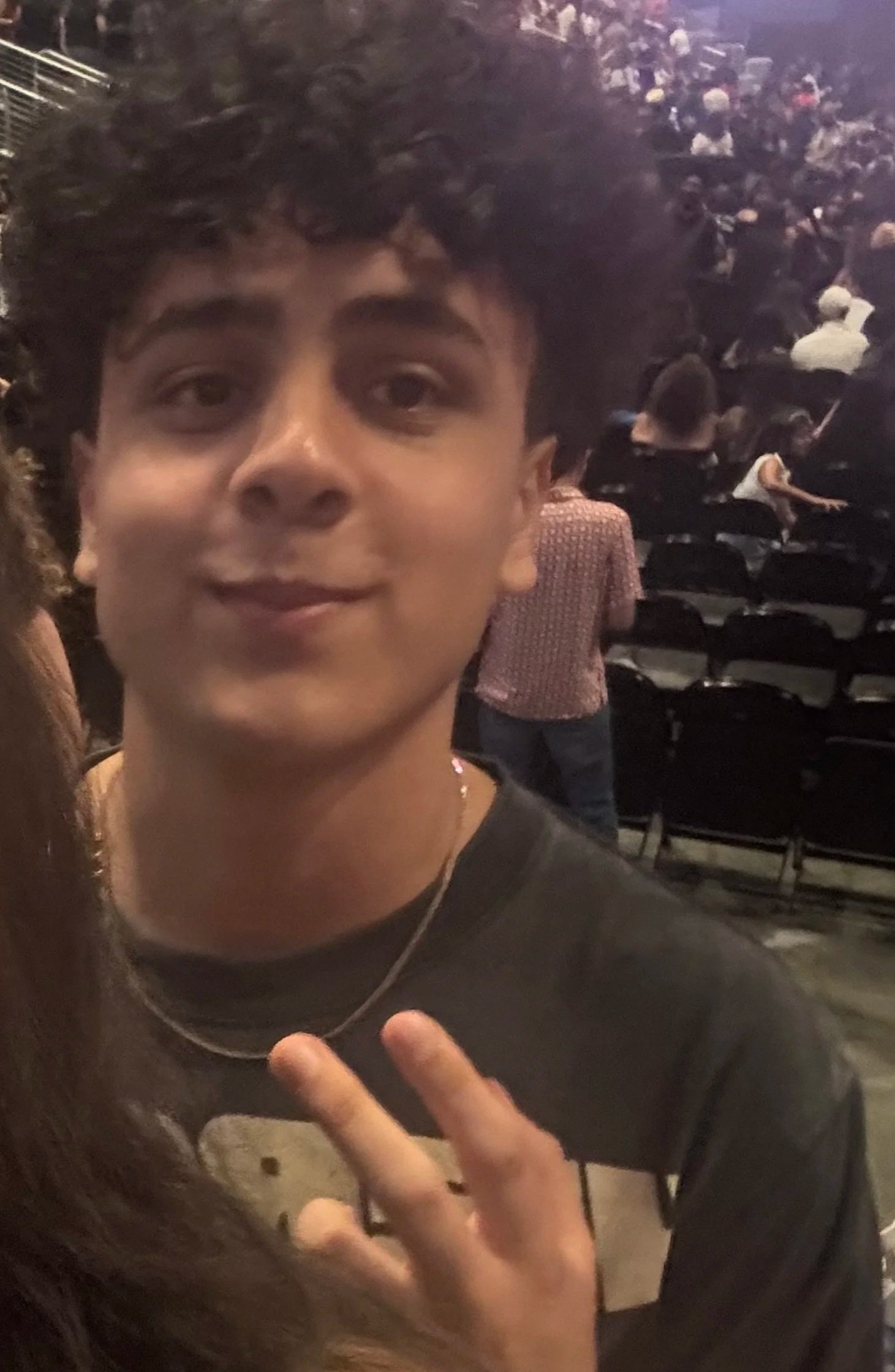 Close-up photo of a young person with curly dark hair making a peace sign with fingers, in a crowded auditorium or theater with rows of seats and many people sitting in the background.