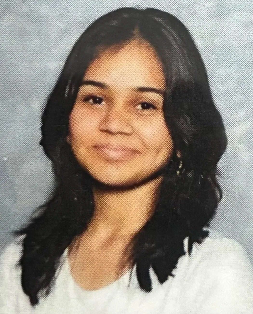 Headshot of a young woman with wavy black hair, smiling and wearing a white top.