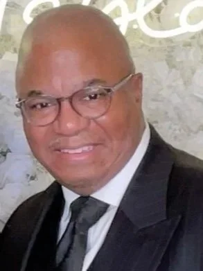Close-up of a smiling man wearing glasses, a black suit, white shirt, and gray tie, with a light-colored background.