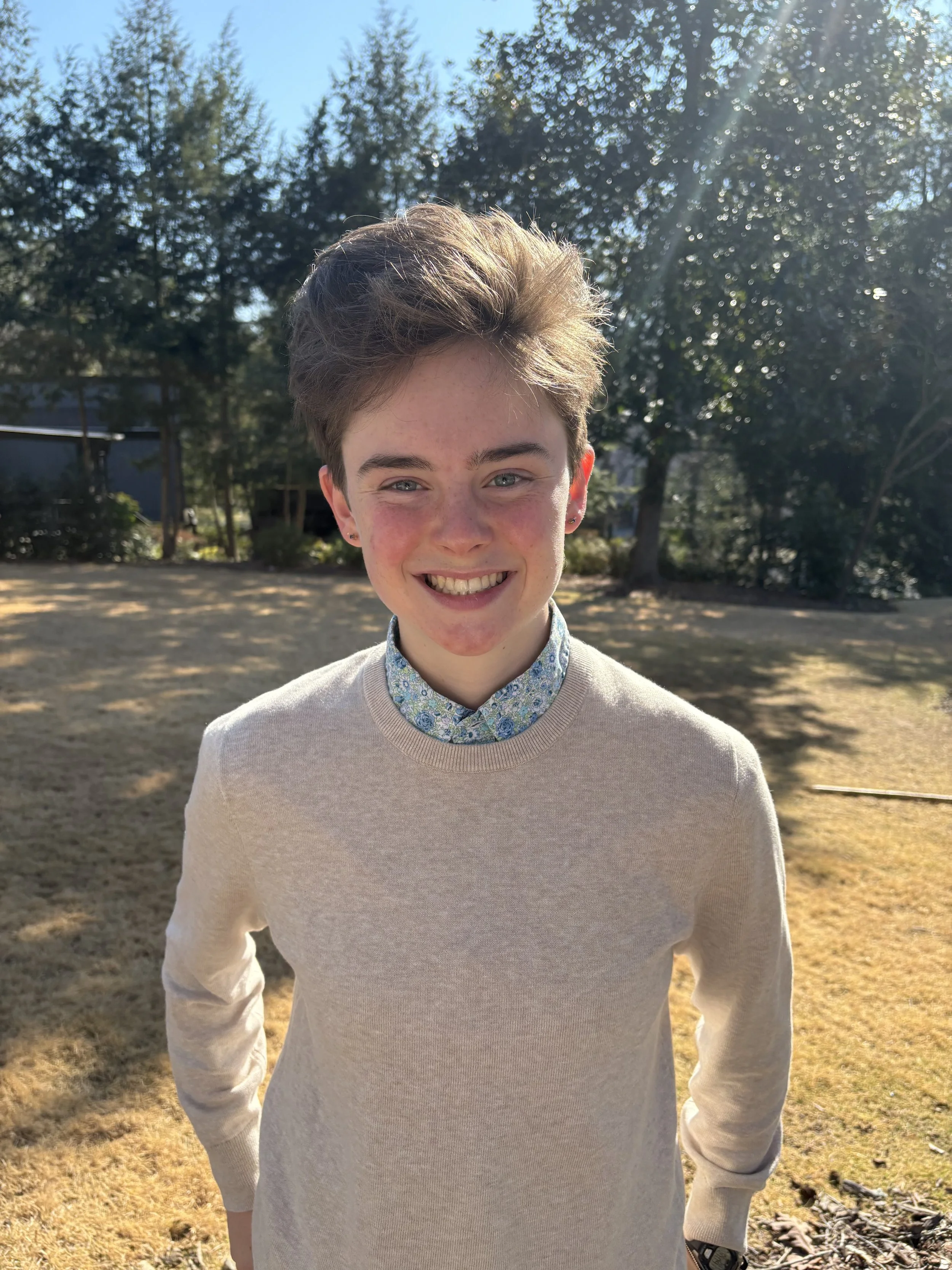 A young person with short, voluminous hair and earrings, smiling outdoors, wearing a light-colored sweater and a patterned collared shirt underneath, with trees and sunlight in the background.