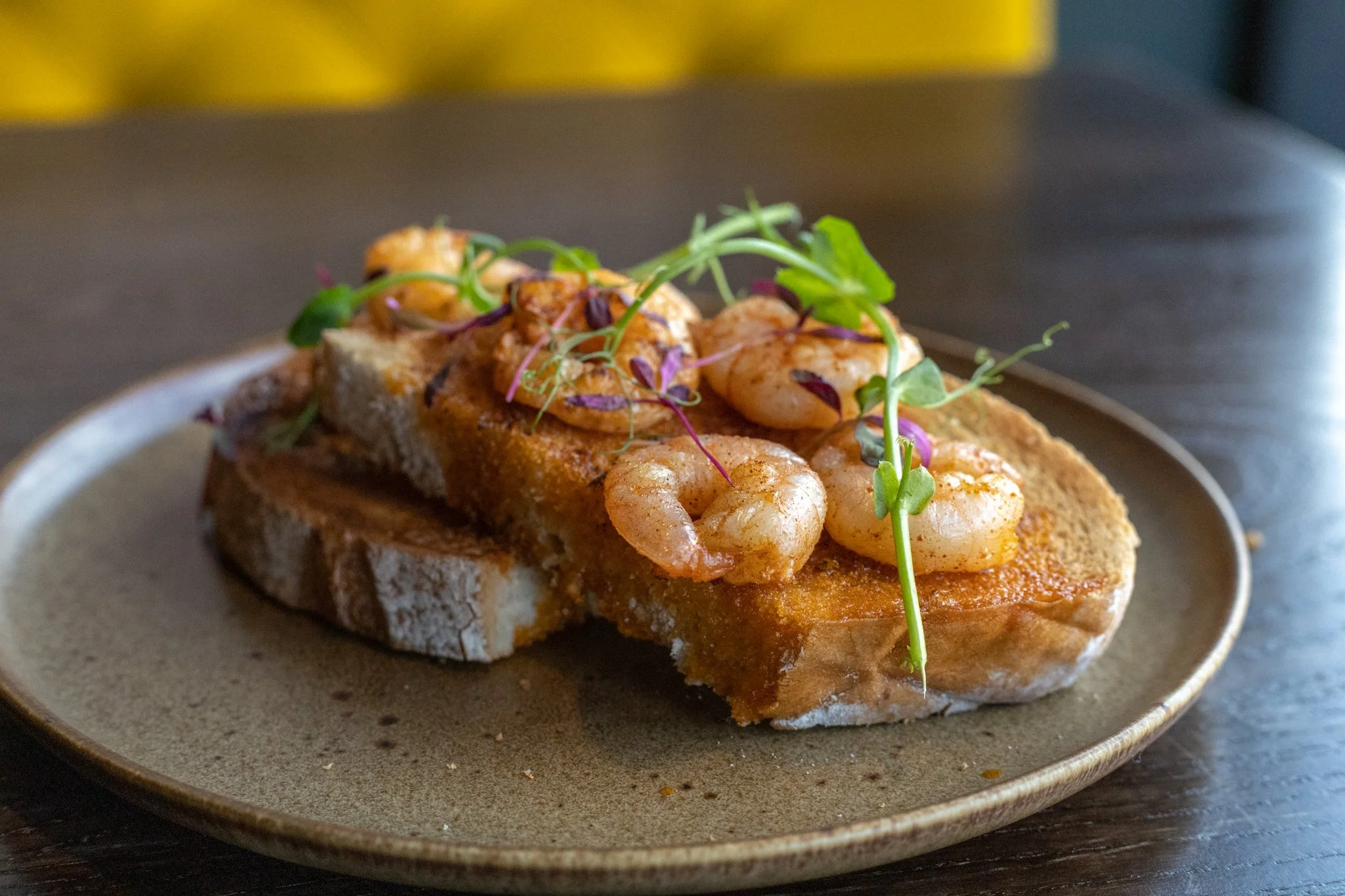 Slice of toasted bread topped with cooked shrimp, microgreens, and garnishes, served on a ceramic plate.