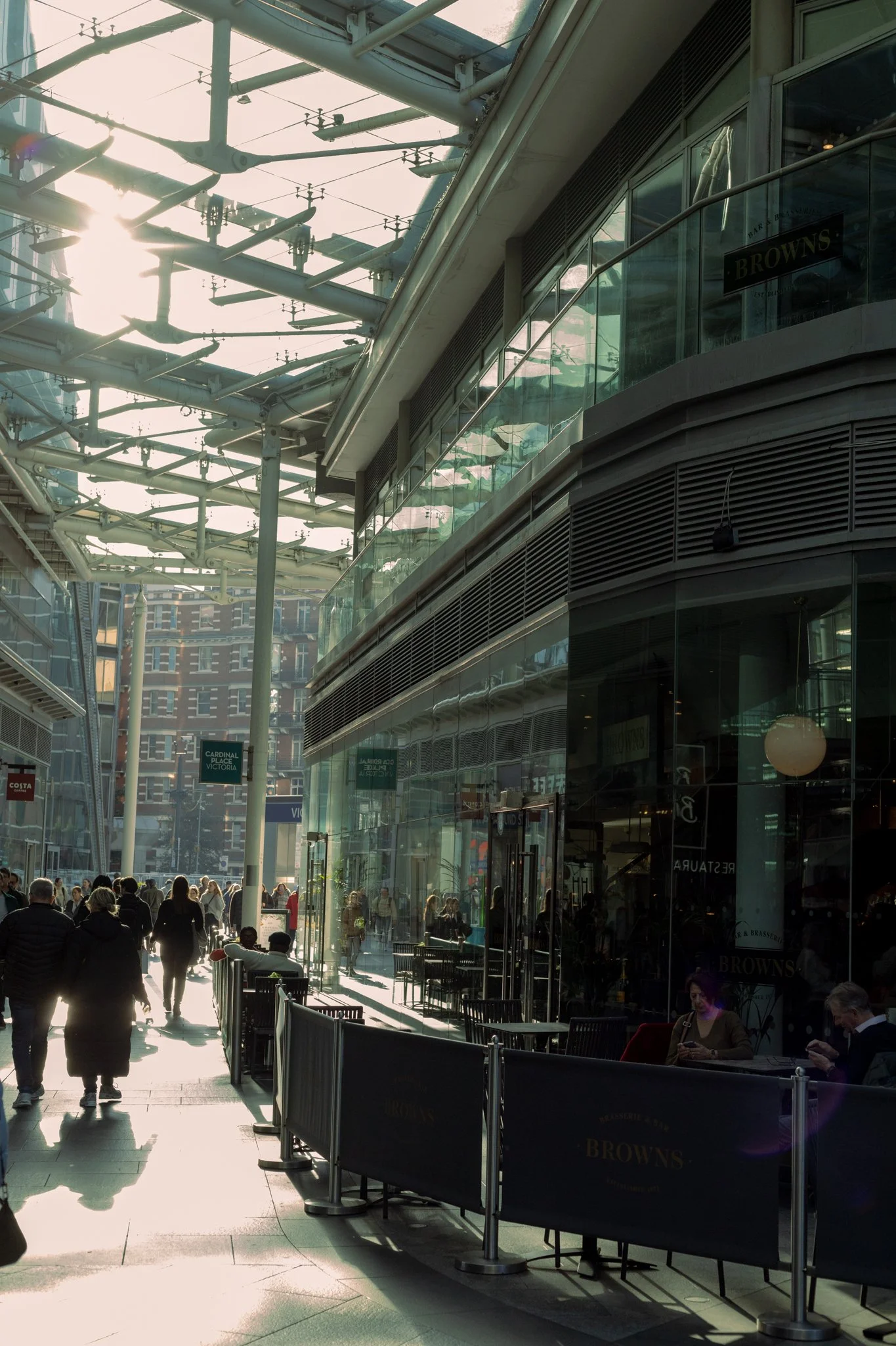 Crowded urban shopping area with people walking and sitting outside a cafe or restaurant, modern glass buildings, and an intricate metal and glass roof structure overhead.
