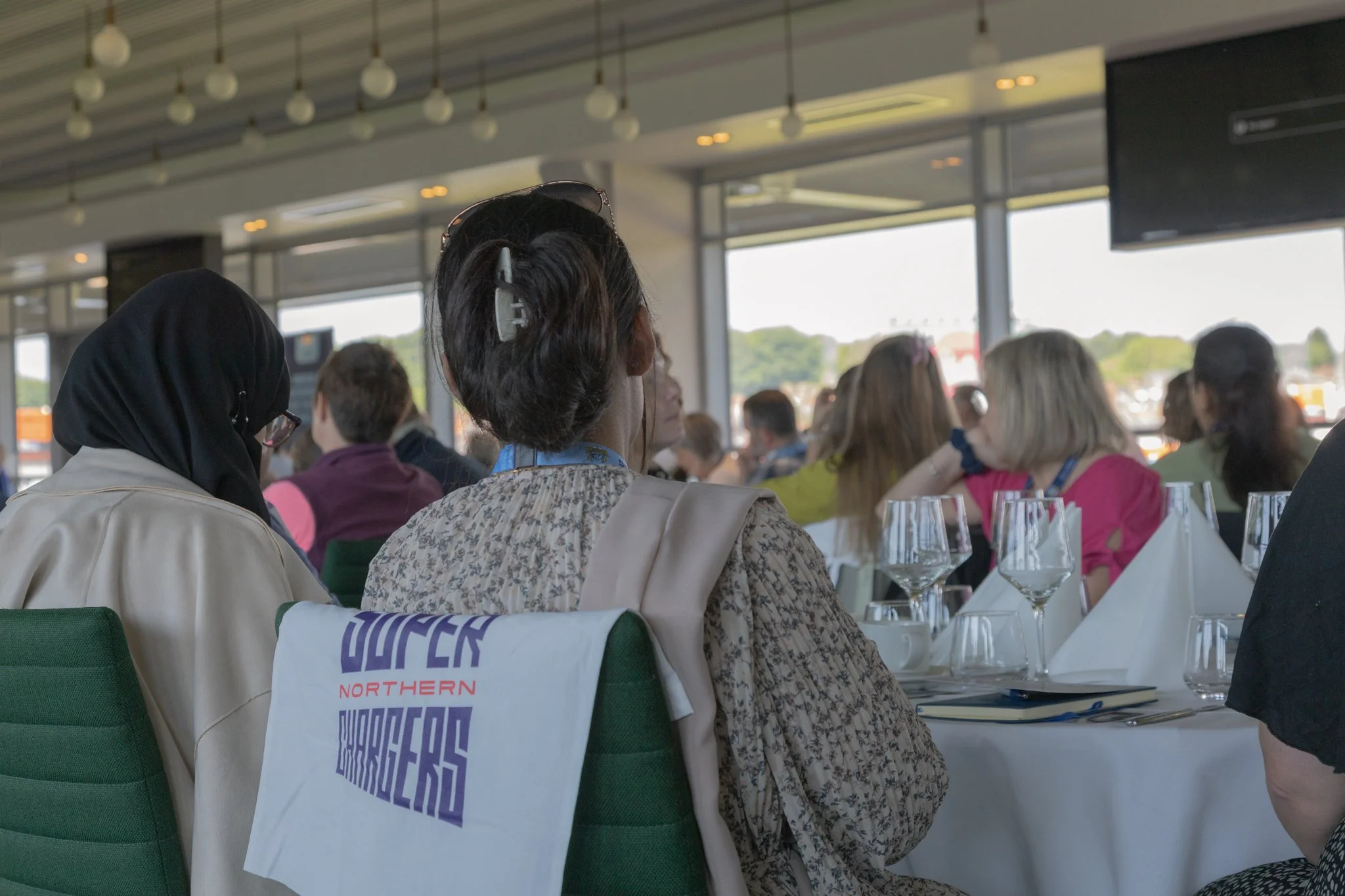 Attendees sitting at tables during a conference or event in a well-lit room with large windows, with place settings including wine glasses and napkins.