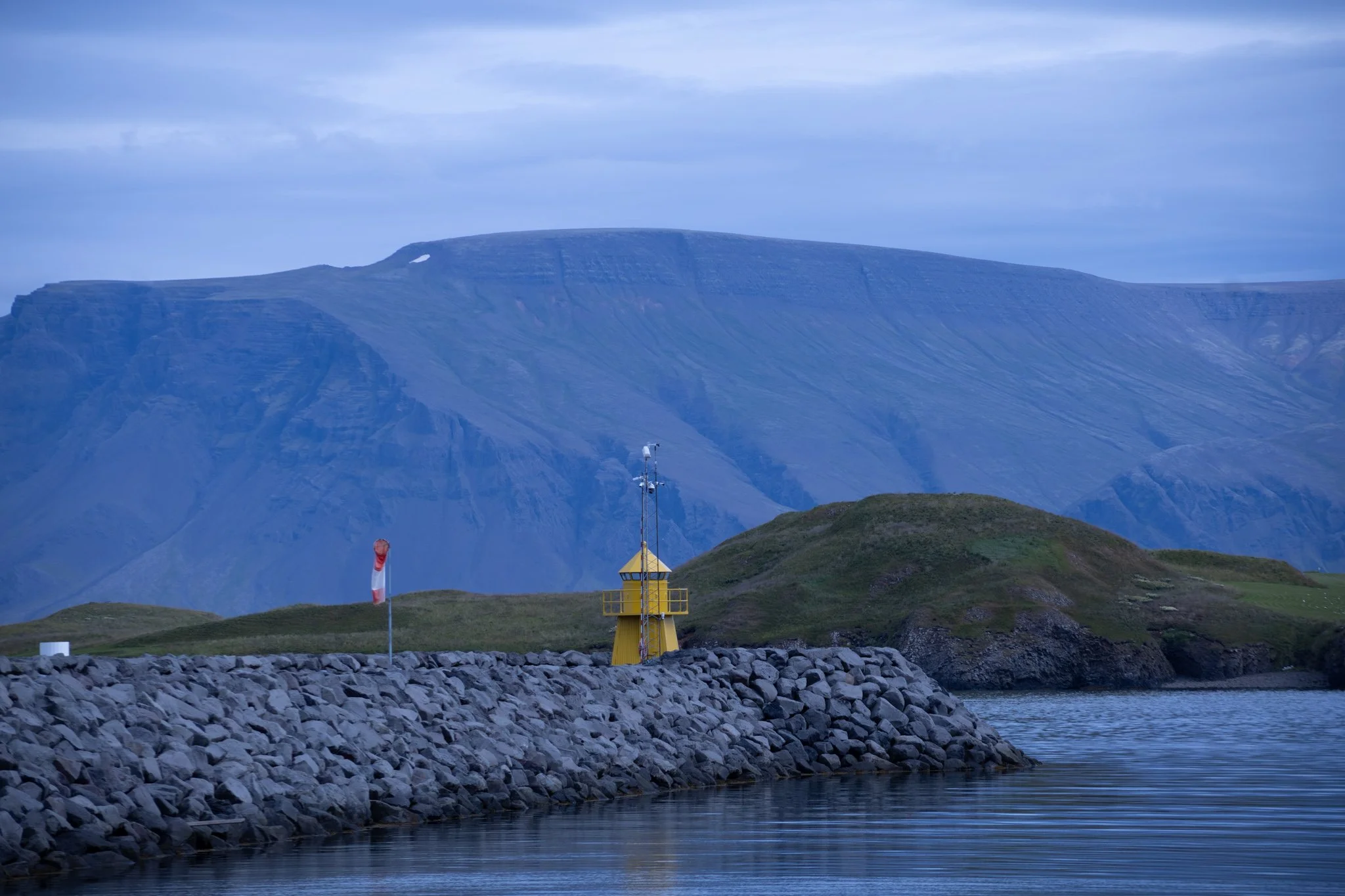 A rocky shoreline with a yellow lighthouse, a hill, and a large mountain in the background under a cloudy sky.
