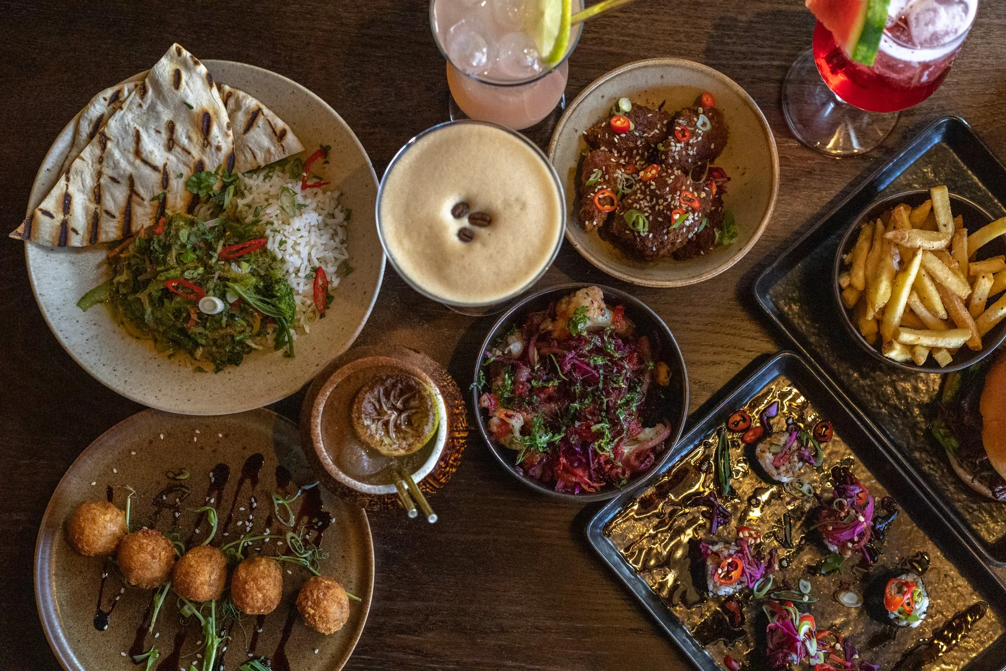 A variety of dishes on a wooden table, including grilled flatbread with rice and vegetables, a glass of pink cocktail with lime, beer with coffee beans on foam, fried chicken bites with chili and green onions, a bowl of salad with purple cabbage, and