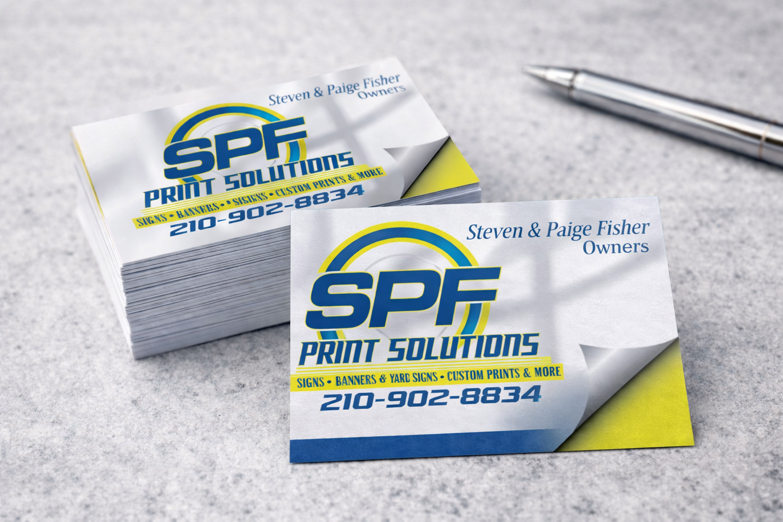 Business cards for SPF Print Solutions with contact number 210-902-8834, stacked on a gray surface with a silver pen nearby.